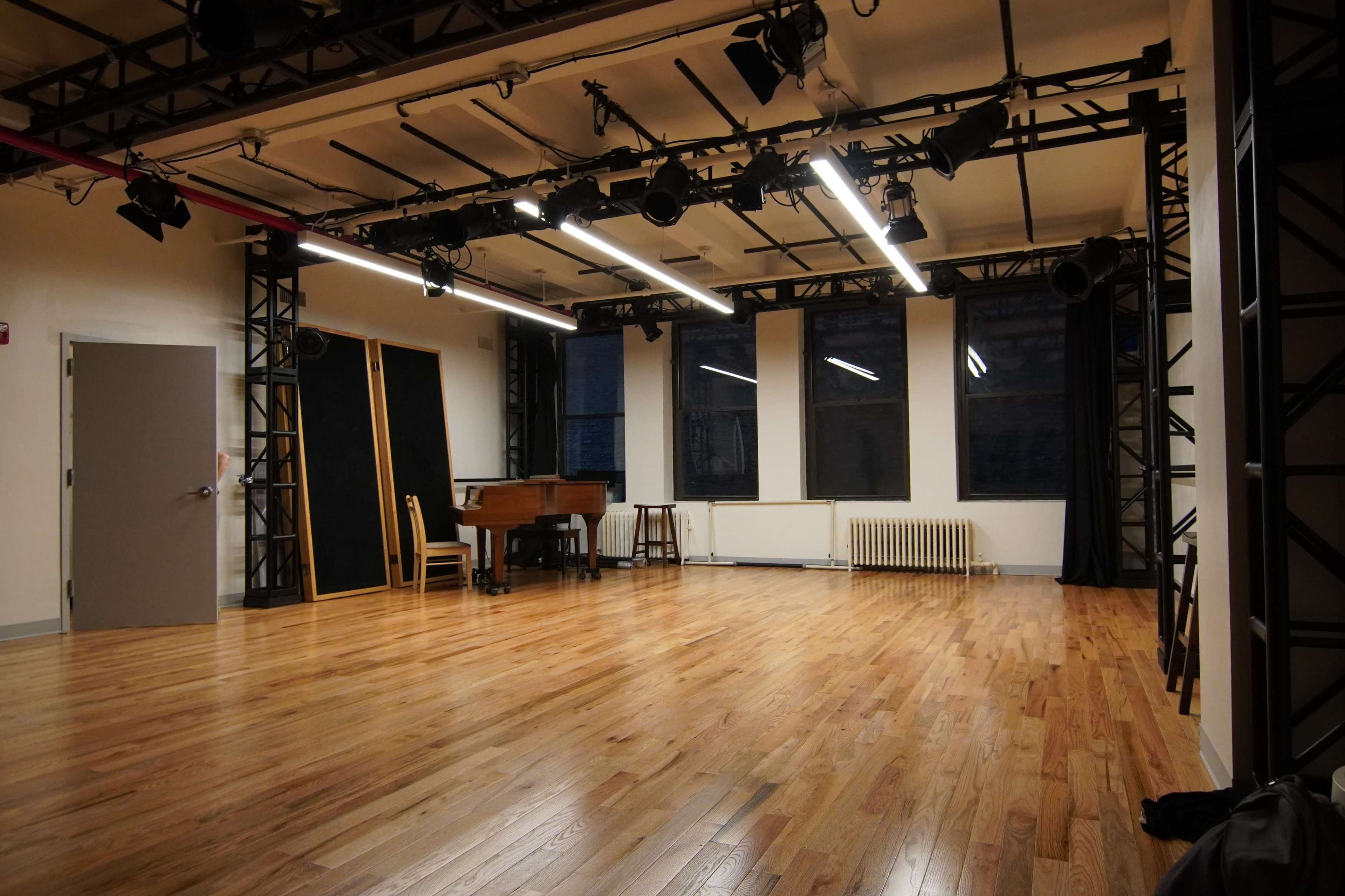 A spacious, empty rehearsal studio with wooden flooring, a grand piano against one wall, and large windows letting in natural light.