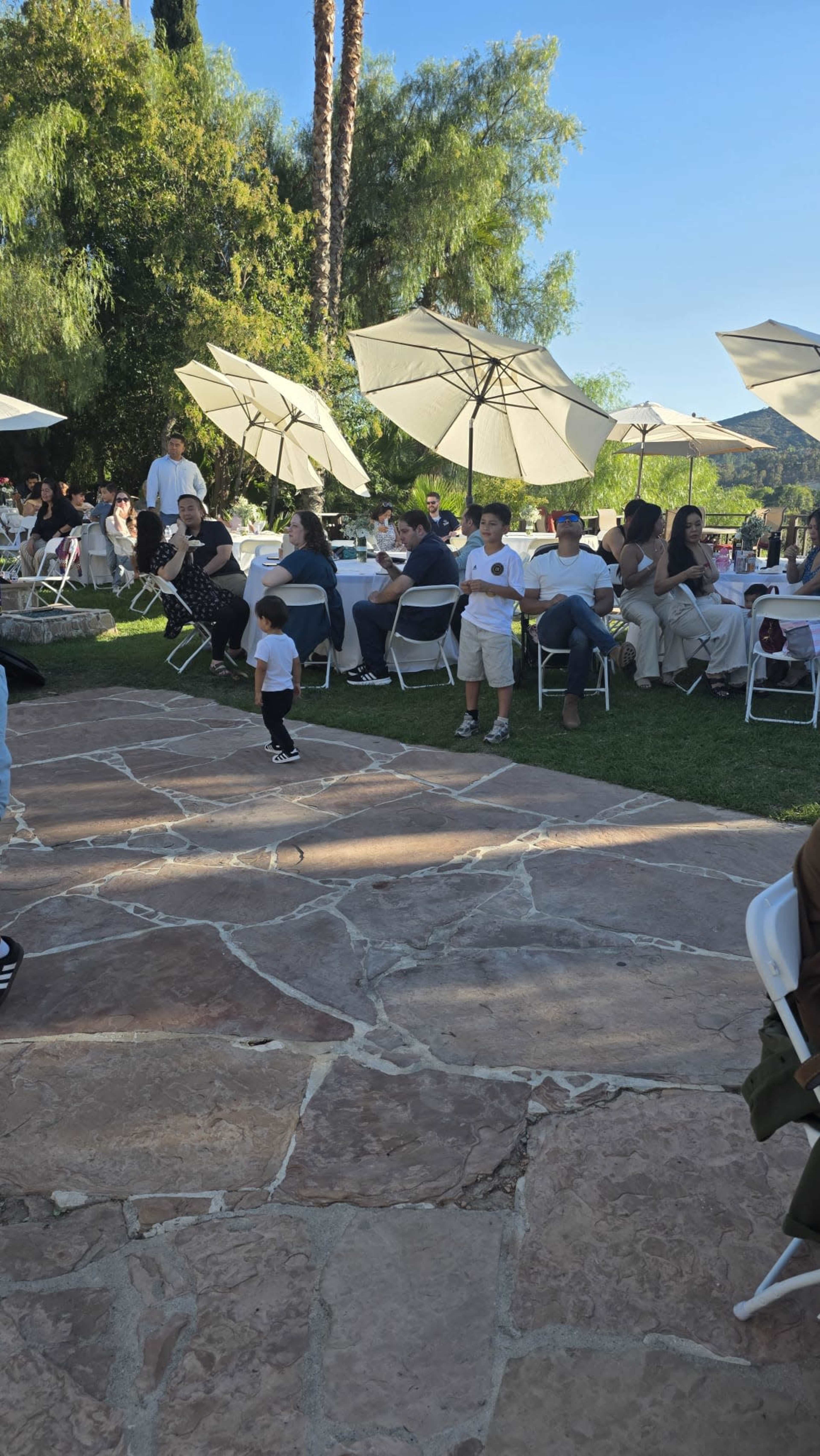 A child in a black outfit dances on a stone pathway while groups of people sit at tables beneath large umbrellas.