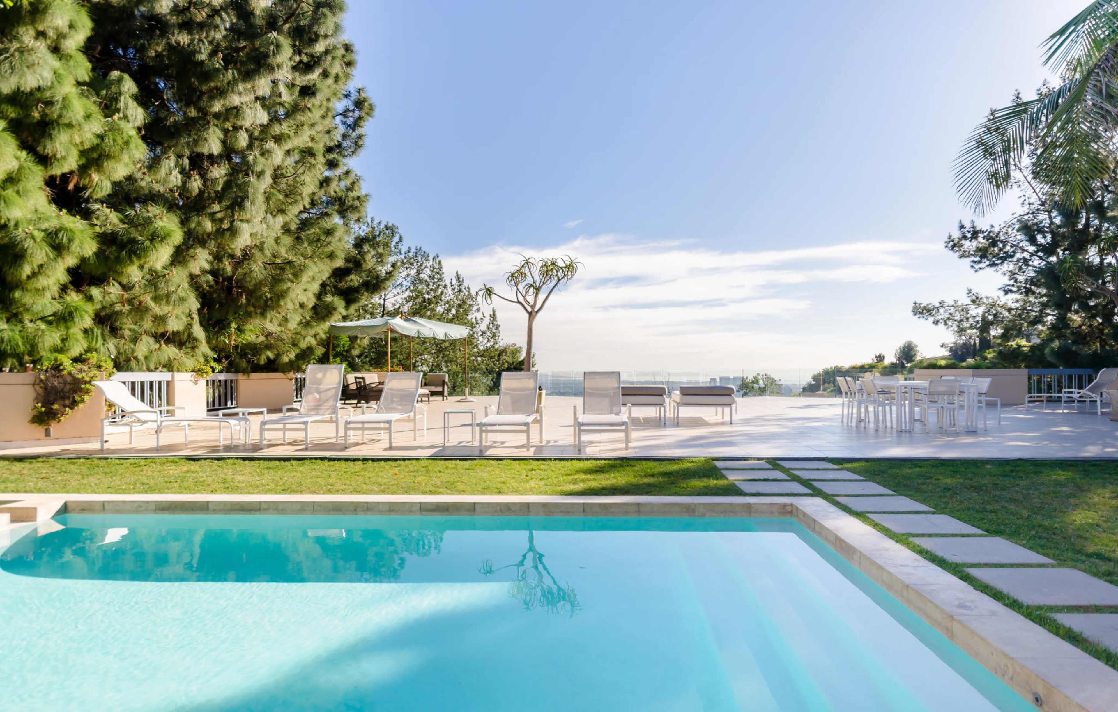 A clear swimming pool surrounded by a lawn and lounge chairs, with a patio and trees in the background under a clear blue sky.