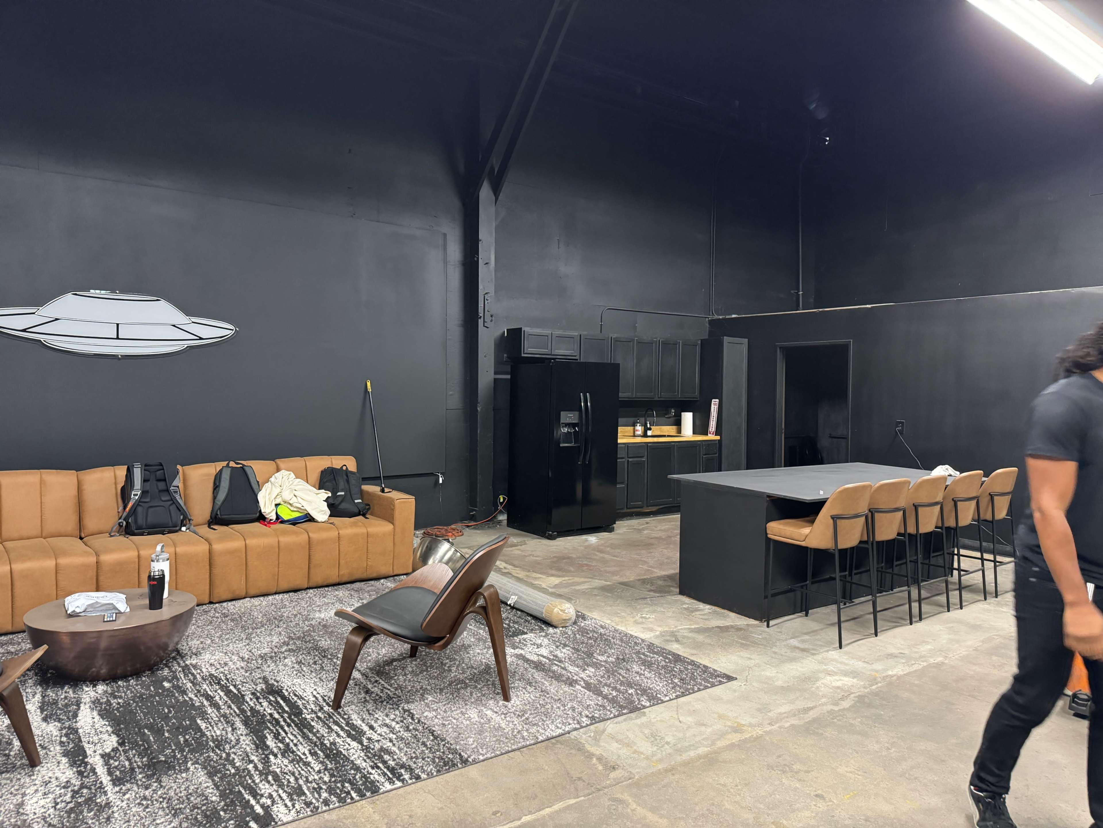 The image shows an industrial-style studio space featuring a brown couch, a black kitchen area with a refrigerator, a dining table with chairs, and a gray area rug on a concrete floor.