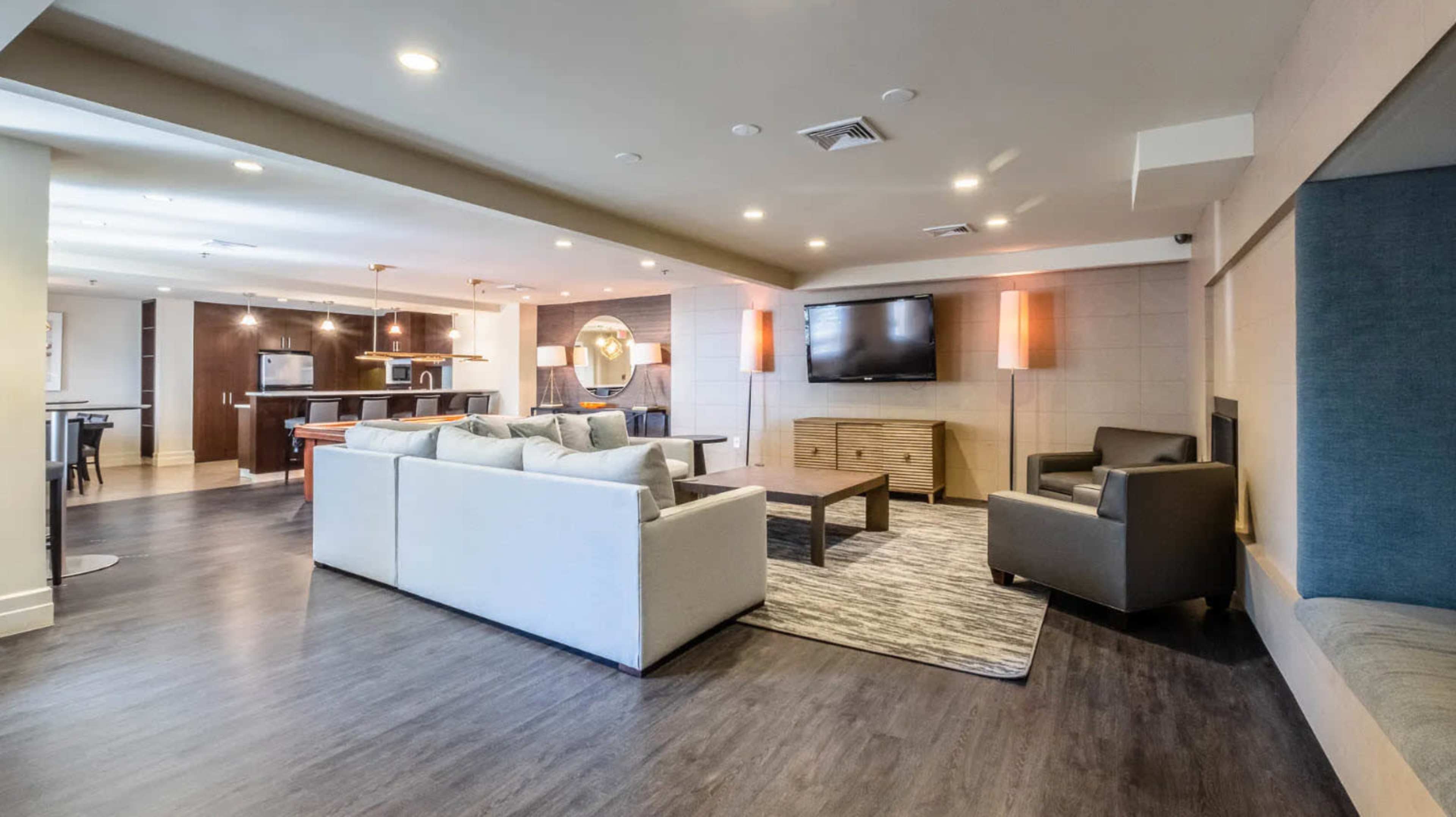 A modern living area with a sofa, two armchairs, a coffee table, and a wall-mounted television, leading into a kitchen space in the background.