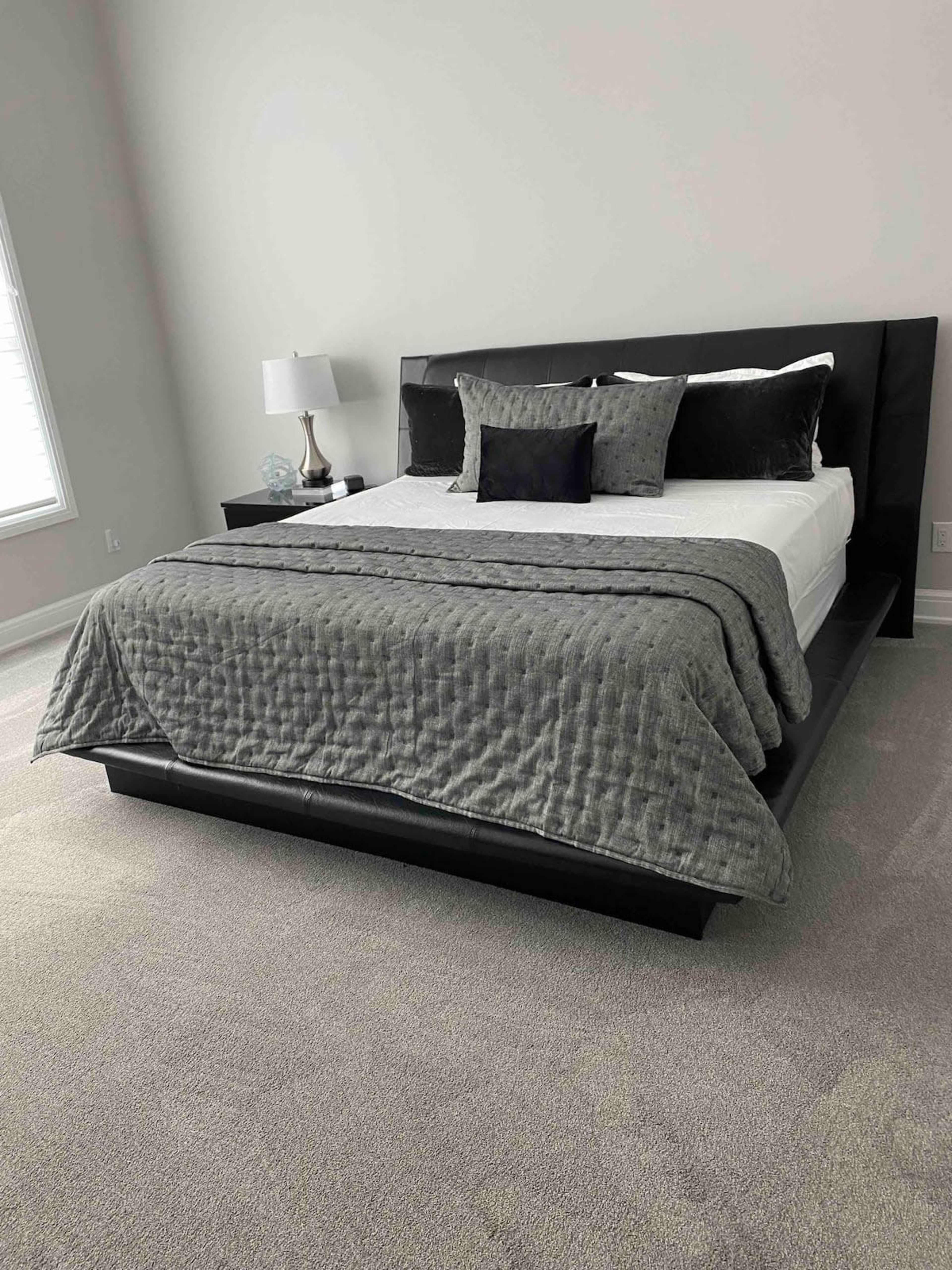 A neatly arranged bed with a dark gray quilt and several pillows is positioned against a light-colored wall in a well-lit bedroom.