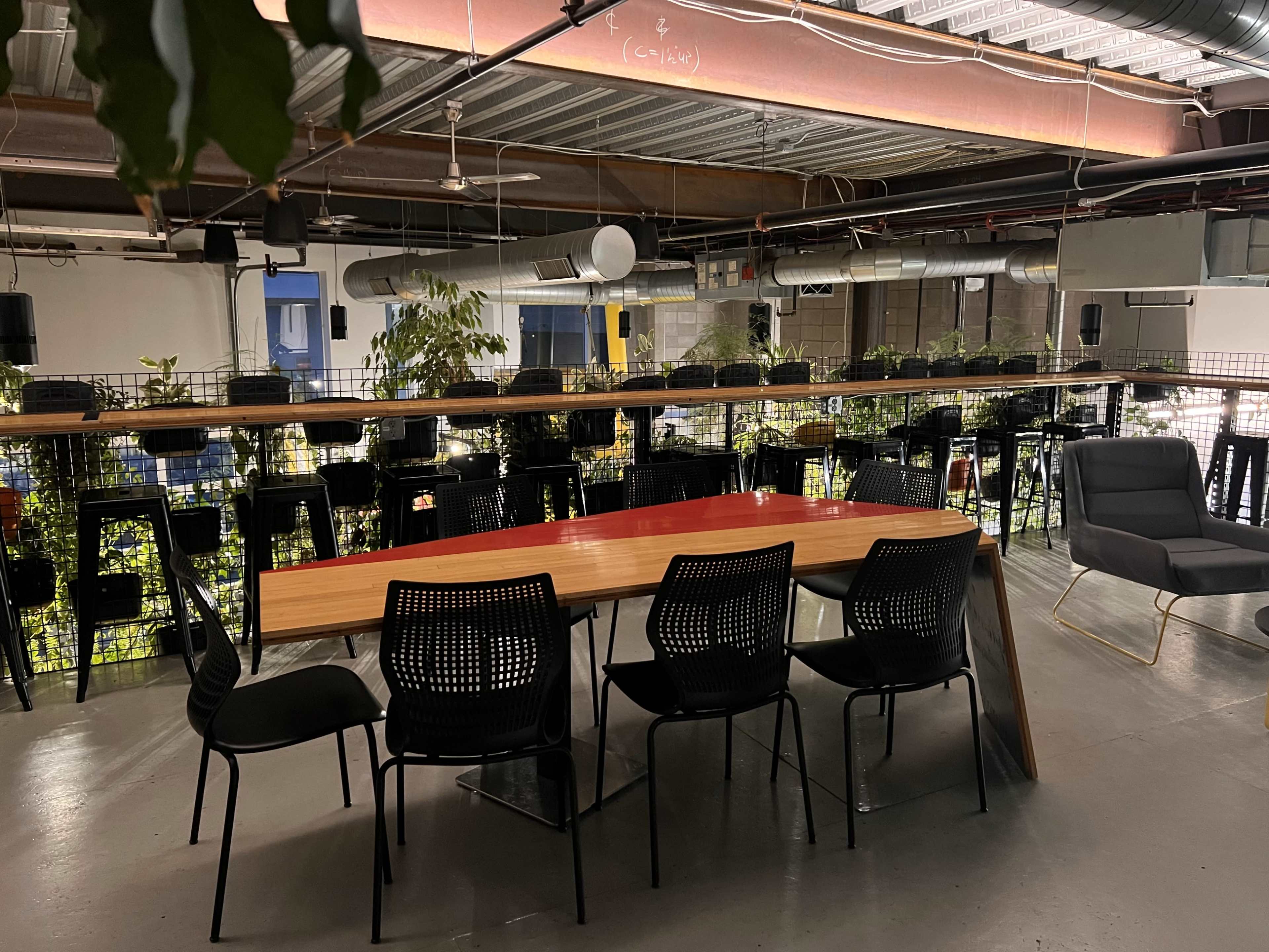 A modern indoor workspace featuring a long wooden table surrounded by black chairs, with plants and high-top seating areas in the background.