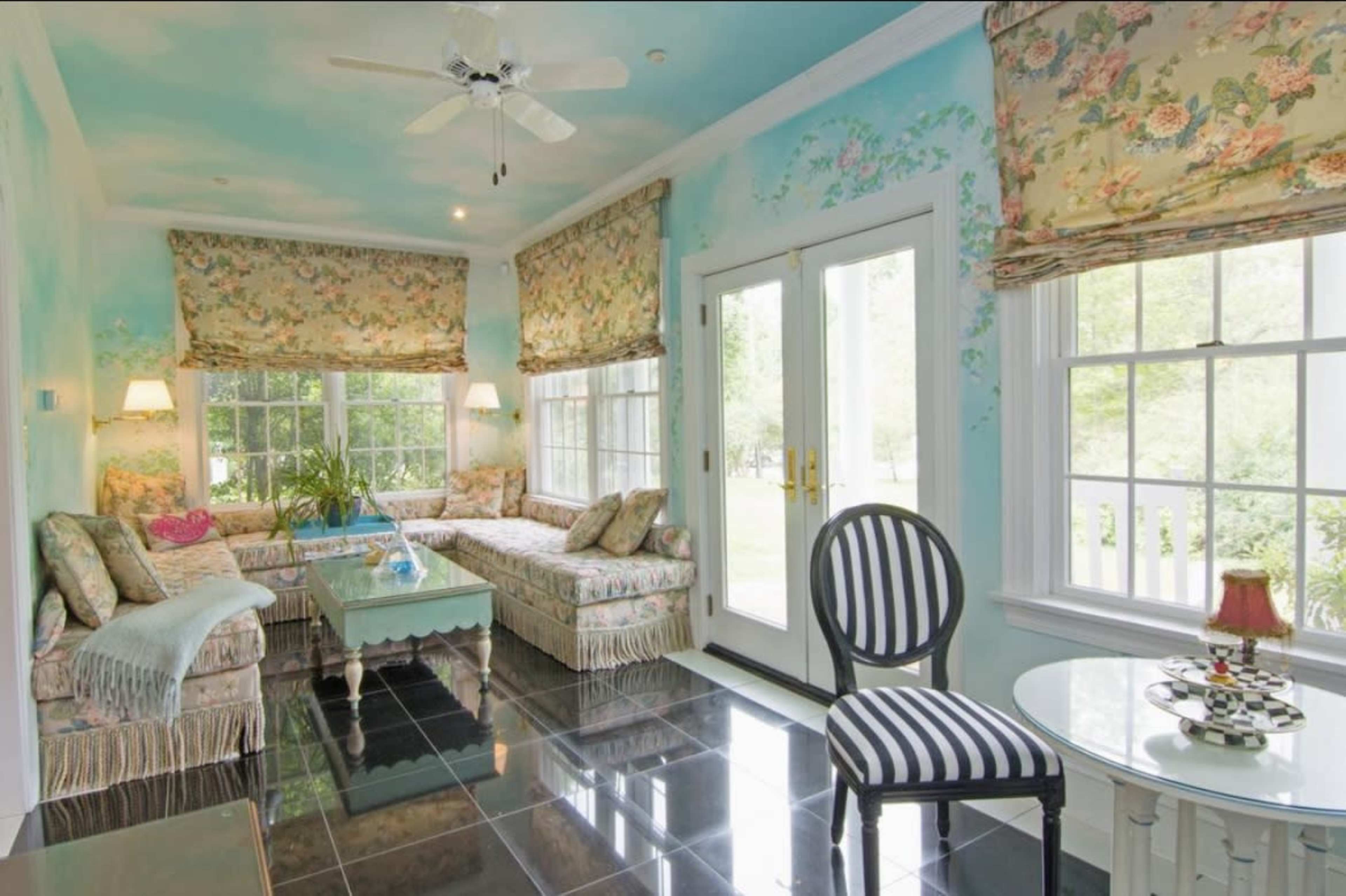 A sunlit sunroom featuring a floral-patterned couch, a round table with a lamp, and large windows with patterned window treatments.