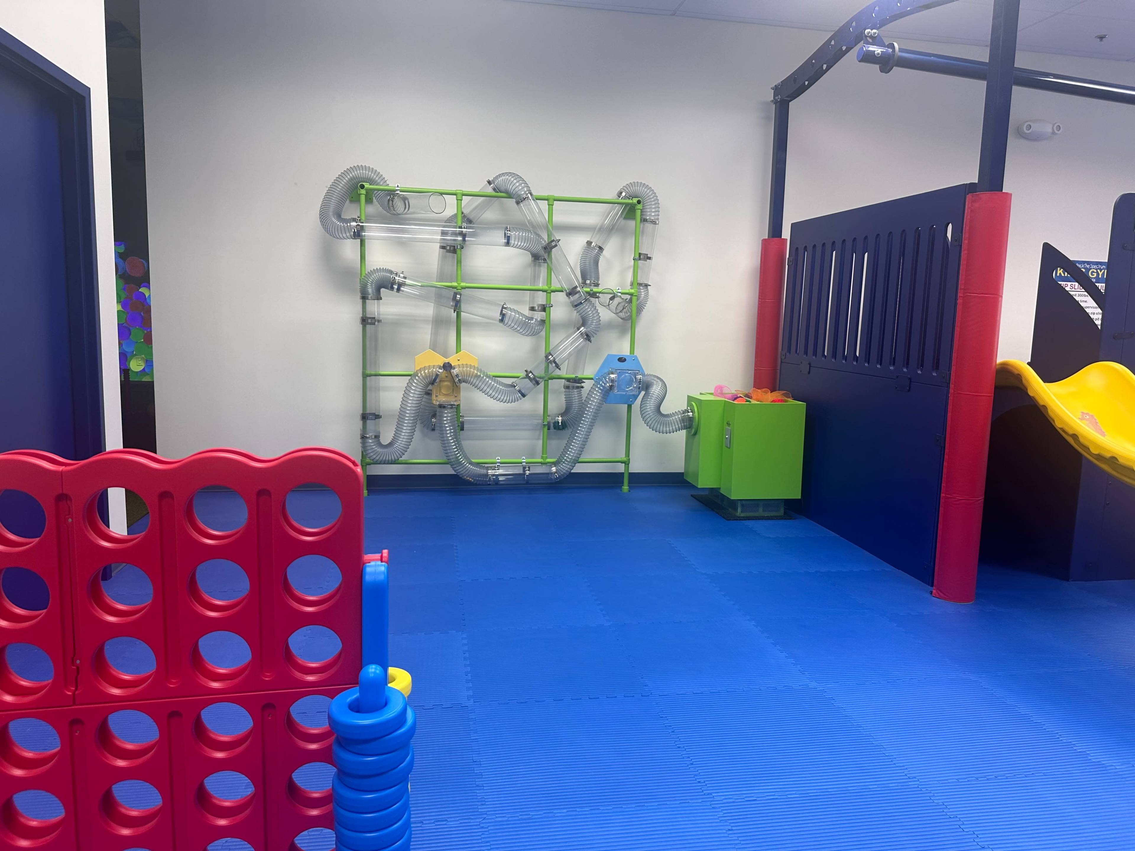 The image shows a colorful indoor play area featuring a green and yellow water play structure, a red connect-four game, and a yellow slide on a blue rubber flooring.
