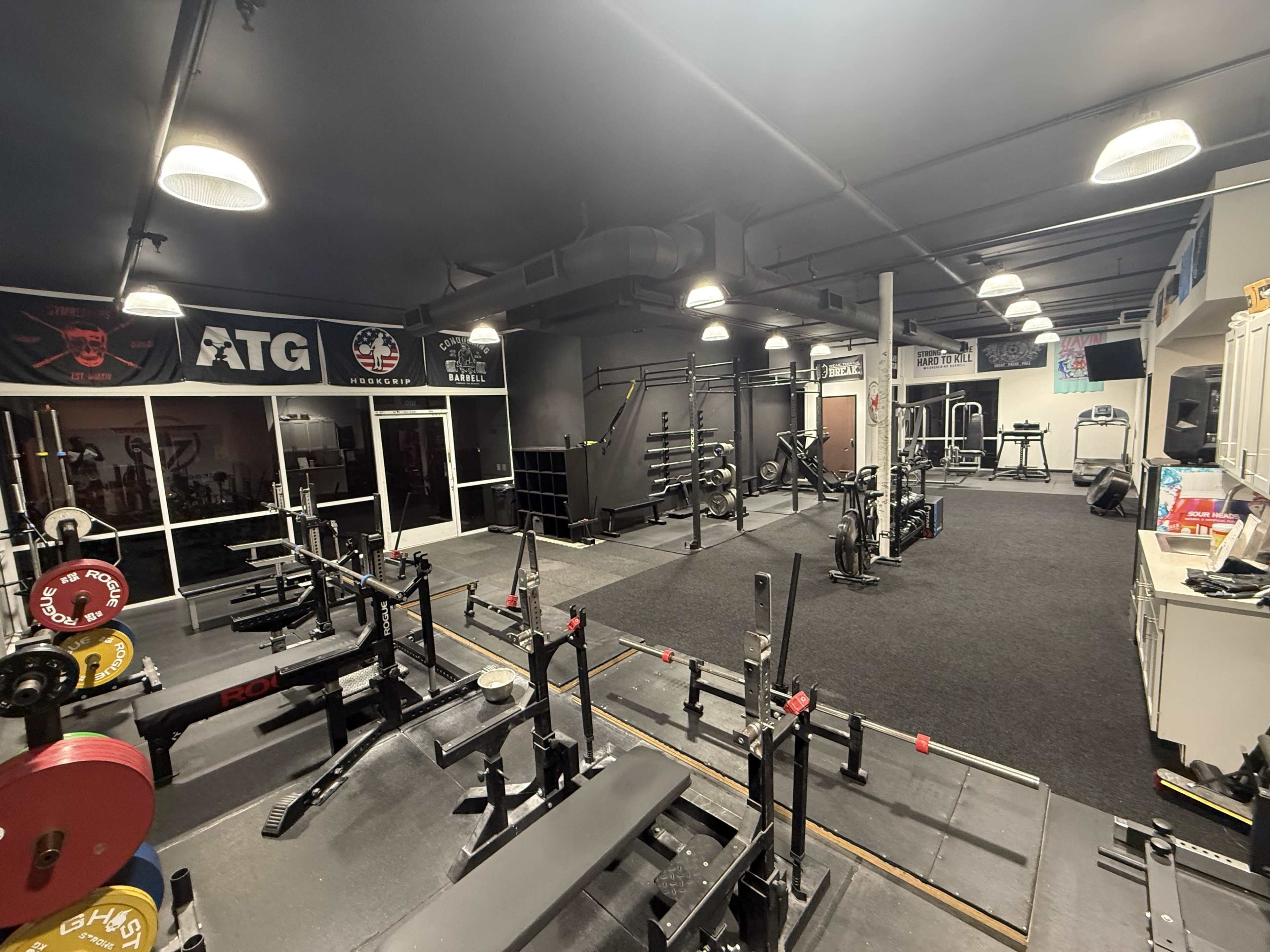 The image shows an indoor gym with various weightlifting equipment, including benches and racks, and well-organized spaces for exercise activities.