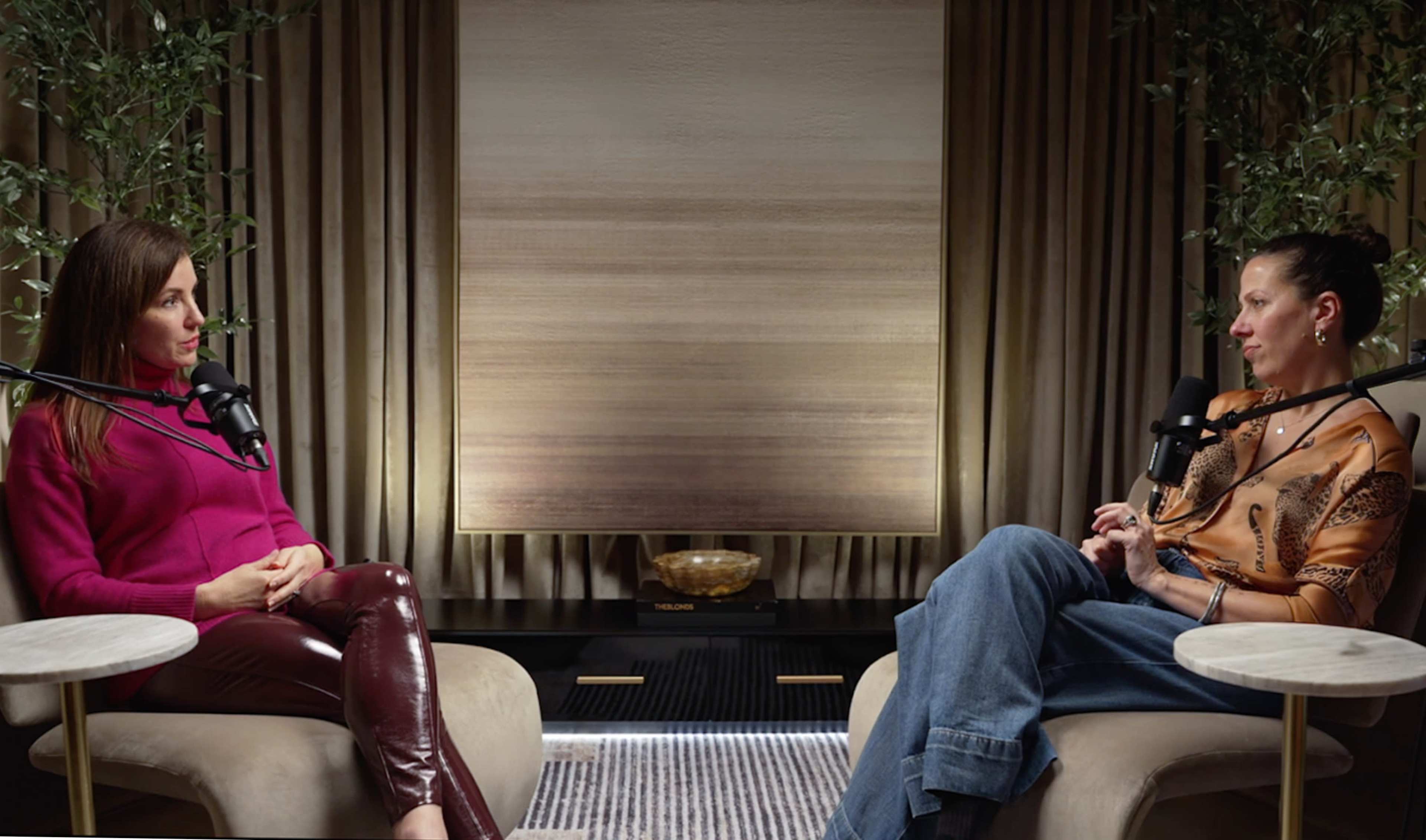 Two women sit in armchairs facing each other in a well-lit, modern interior, with a large abstract painting hanging on the wall behind them.