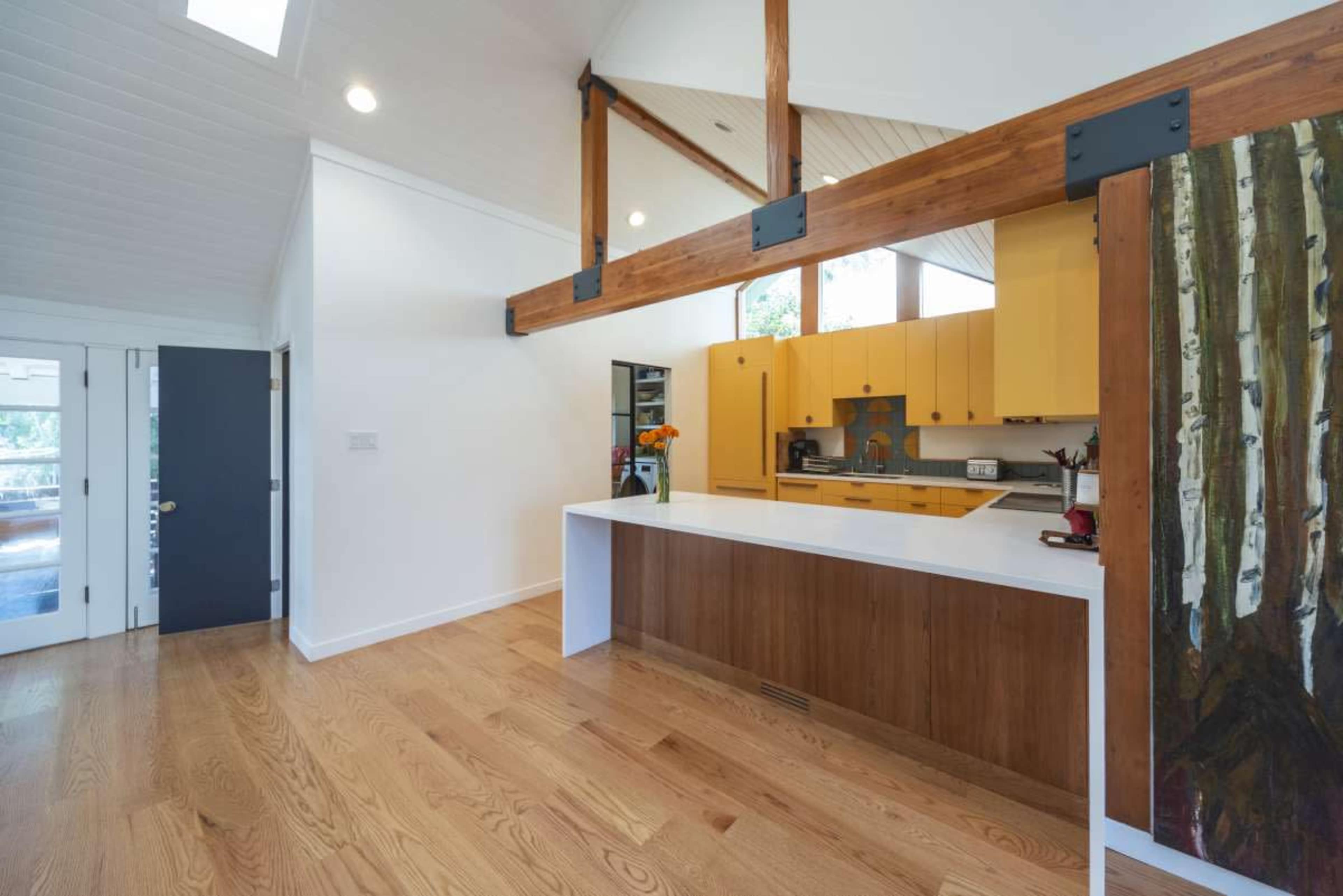 The image shows a modern kitchen with yellow cabinets, a white countertop, and wooden accents, featuring large windows and an open layout.