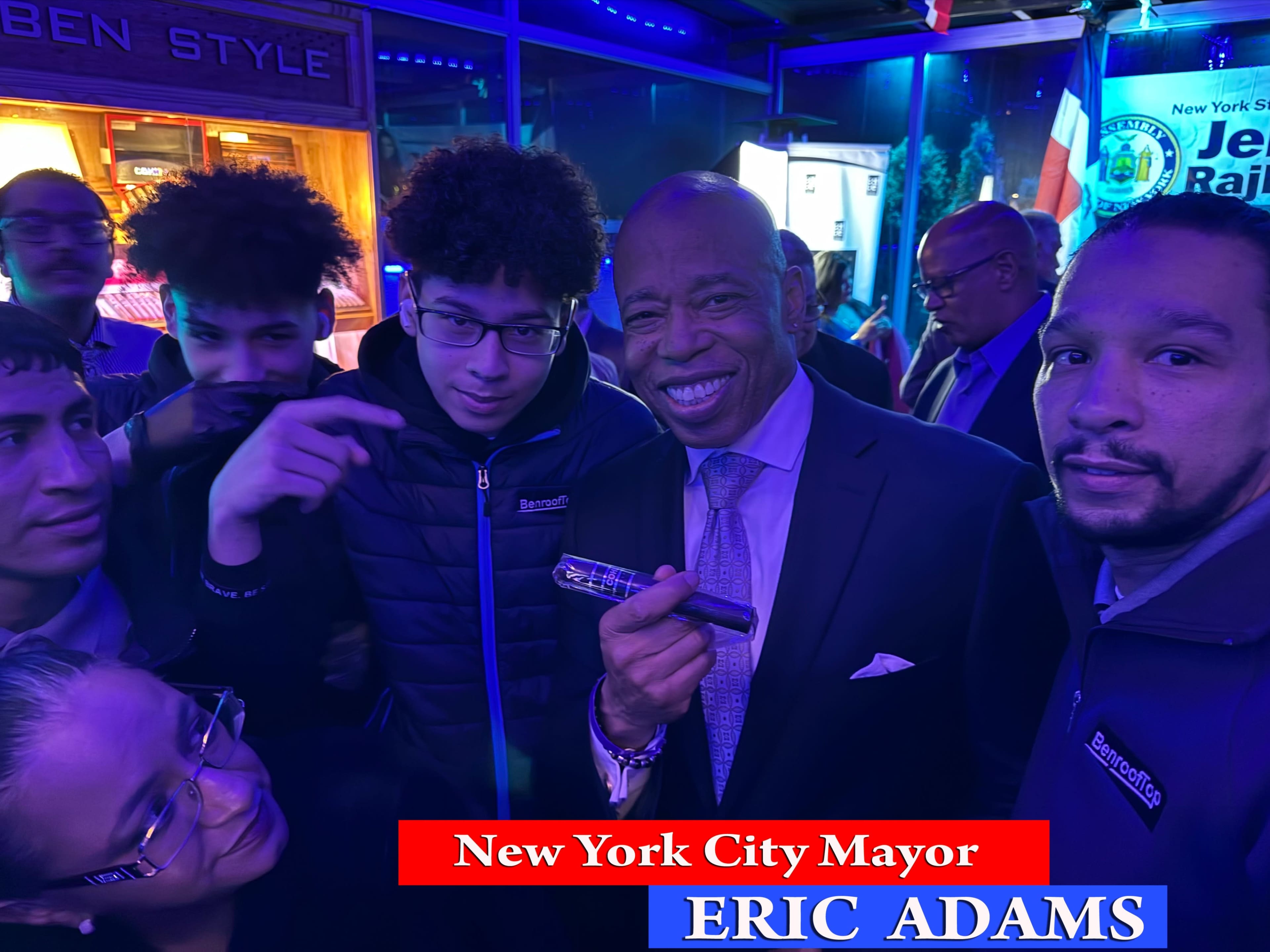 A group of people gathers around New York City Mayor Eric Adams, who is smiling and holding a microphone in a dimly lit venue.