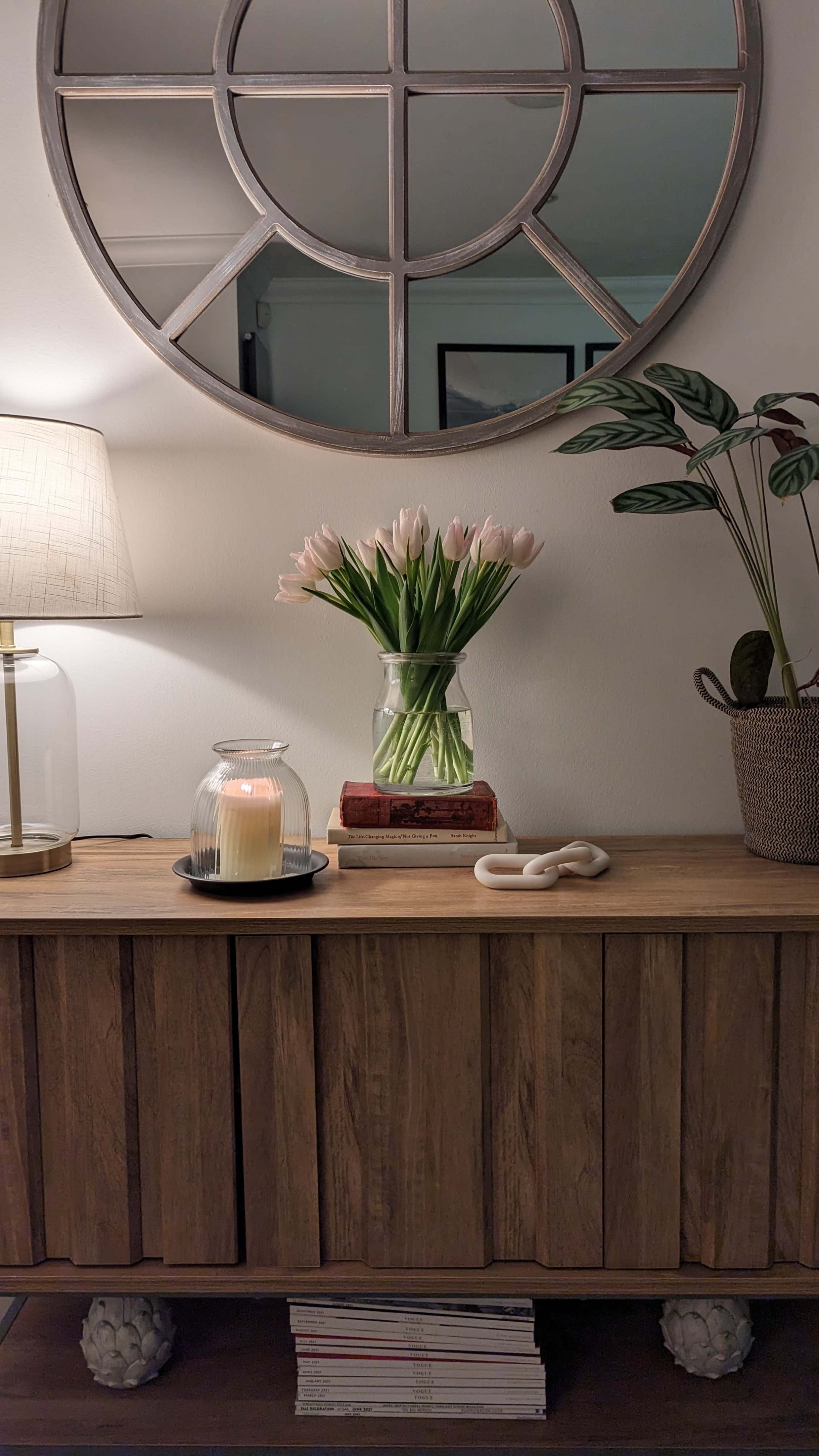 A wooden sideboard displays a vase of pink tulips, a lit candle in a glass holder, a lamp, and decorative items, with a large round mirror reflected on the wall above.
