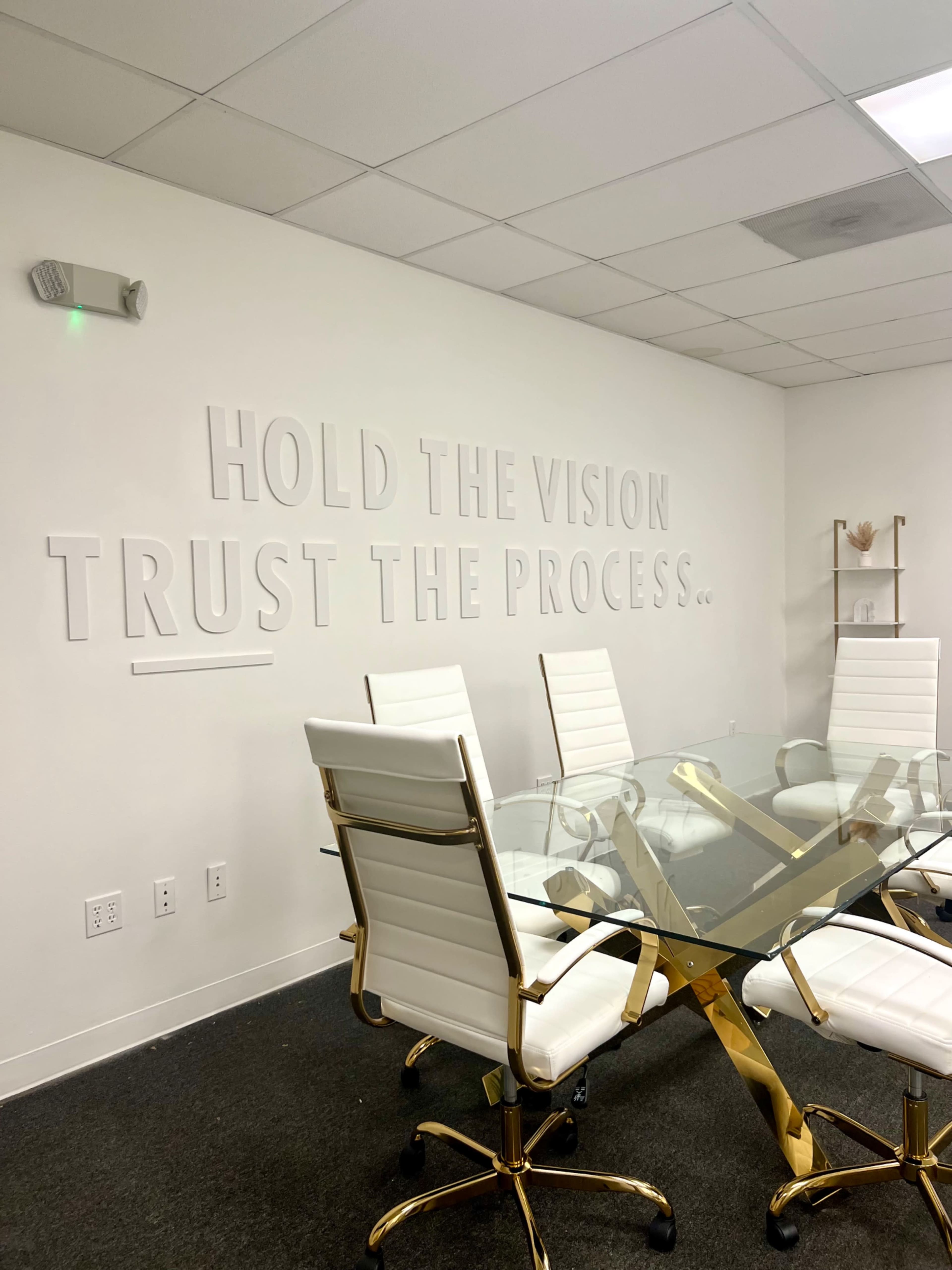 The image shows a modern conference room featuring a glass table surrounded by white chairs, with motivational text on the wall that reads "HOLD THE VISION TRUST THE PROCESS."