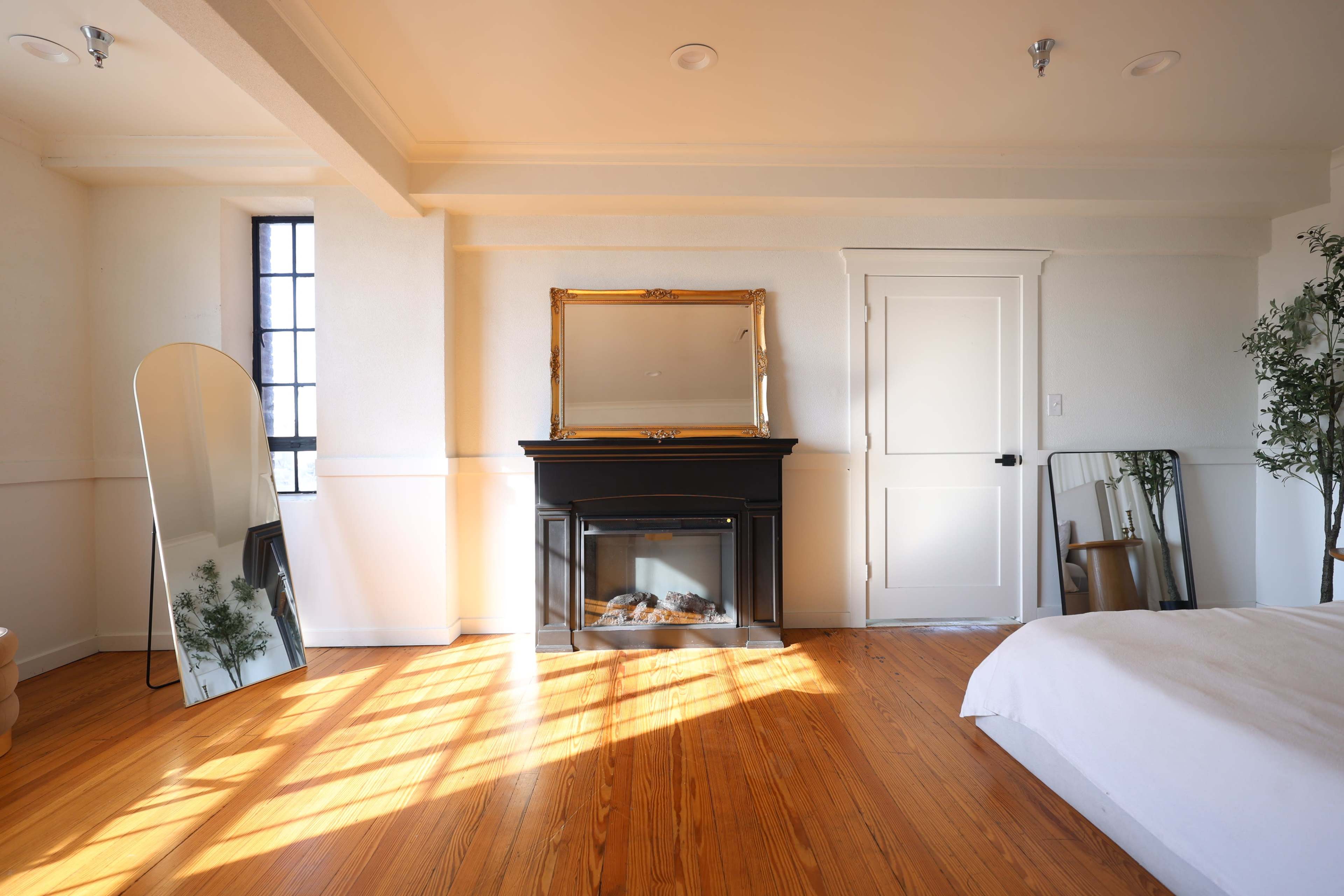 A bright bedroom features a black fireplace, a gold-framed mirror above it, and large mirrors leaning against the walls, with sunlight casting shadows on the wooden floor.