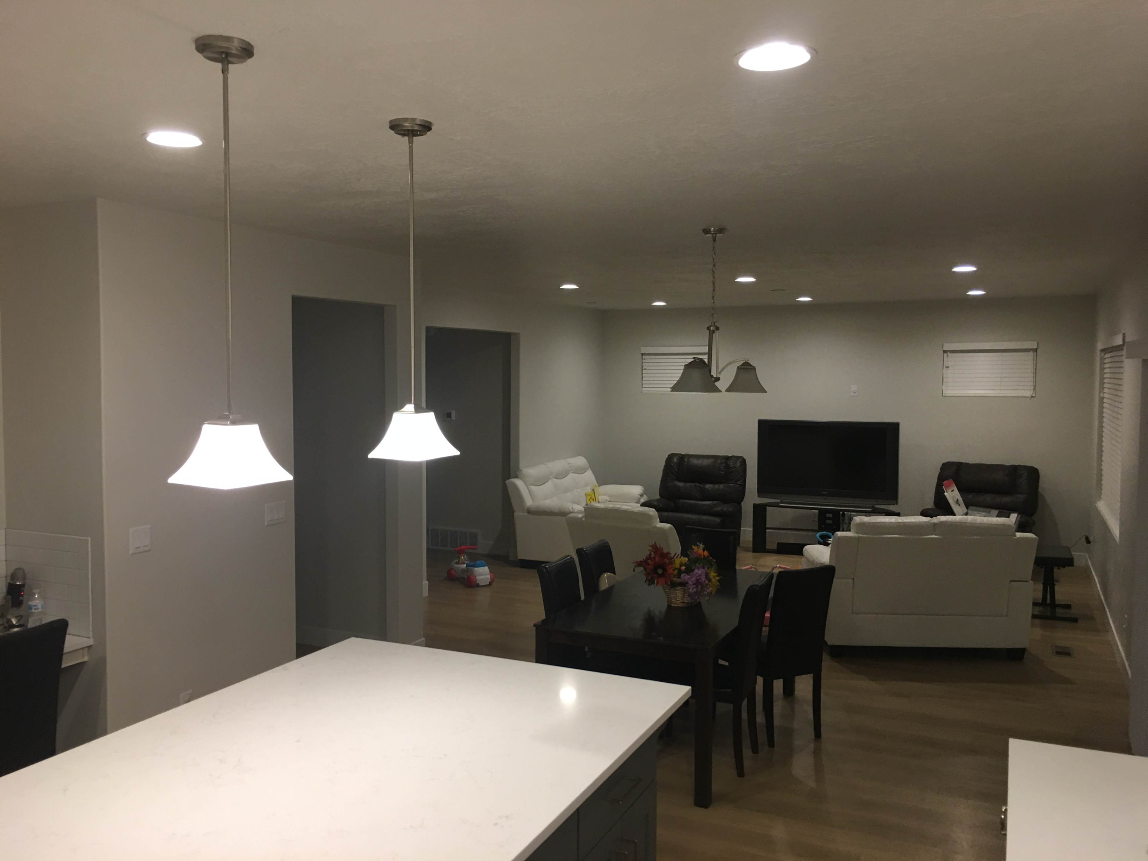 The image shows a modern living room with white walls, light-colored furniture, and a flat-screen TV, illuminated by pendant lights above a kitchen island.