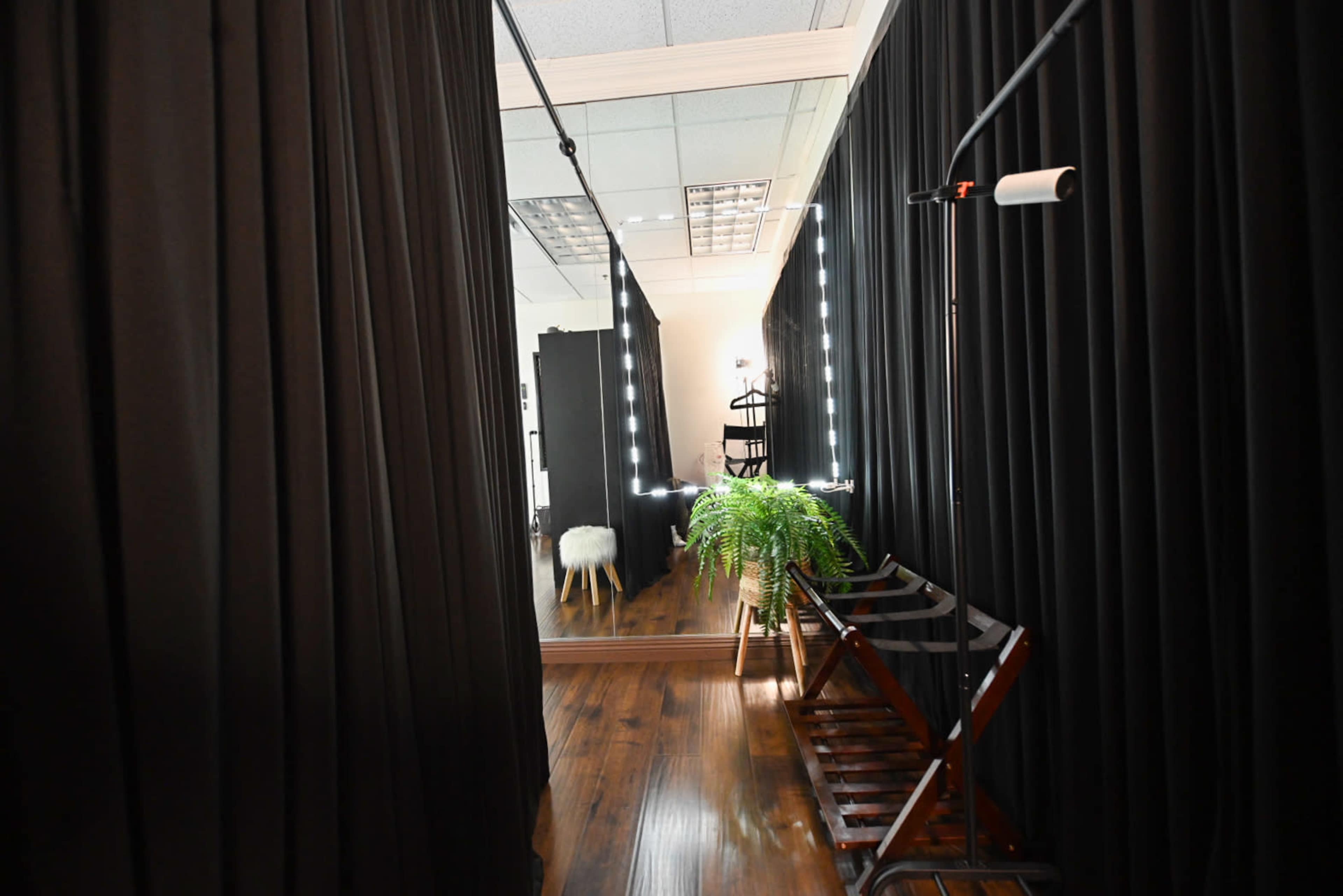 The image shows a narrow hallway lined with black curtains, leading to a mirrored wall with bright lights and a potted plant.