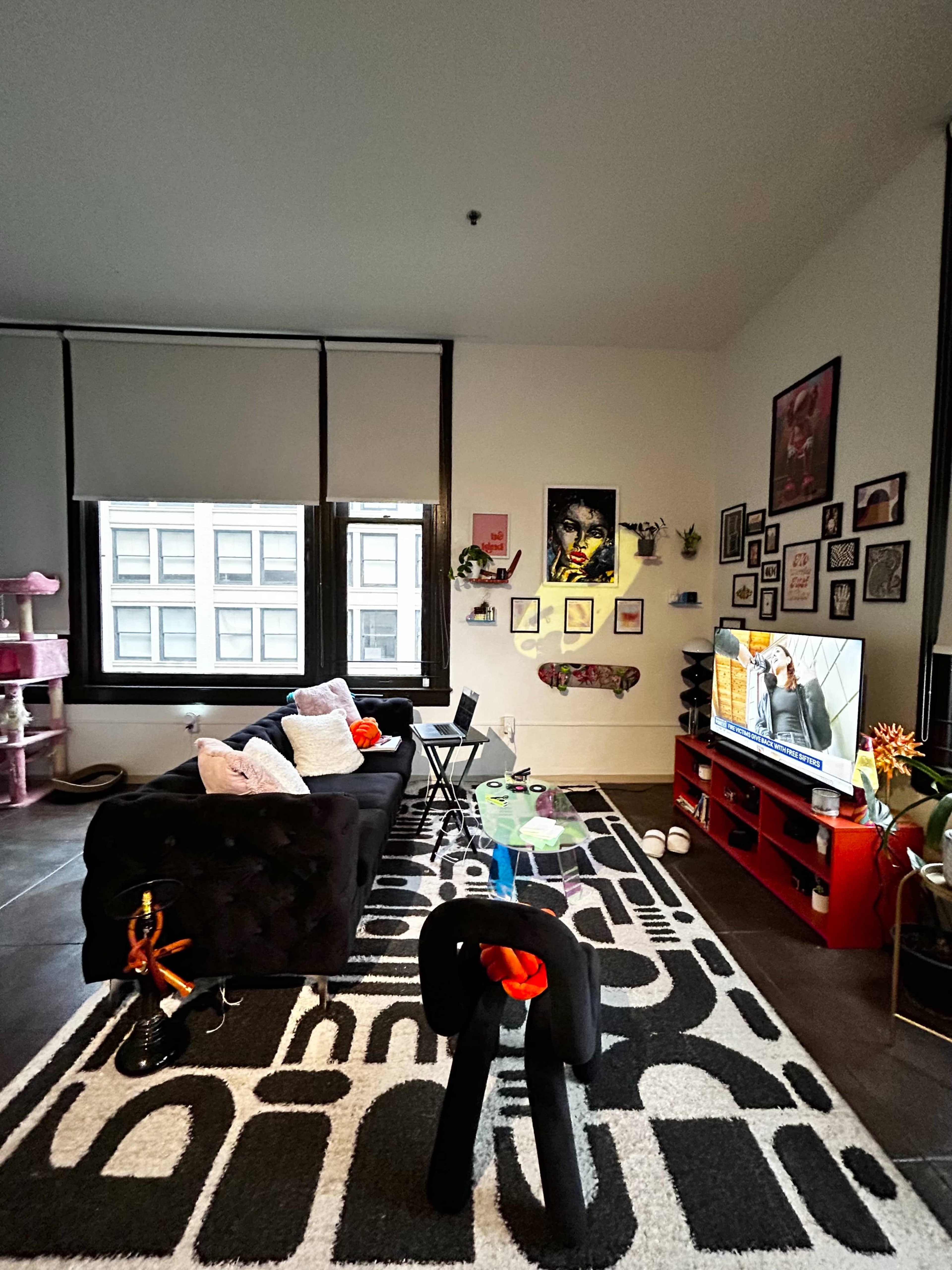 The image shows a modern living room with a black sofa, a television on a red media console, and an array of framed pictures on the walls, along with a patterned rug and various decorative items.
