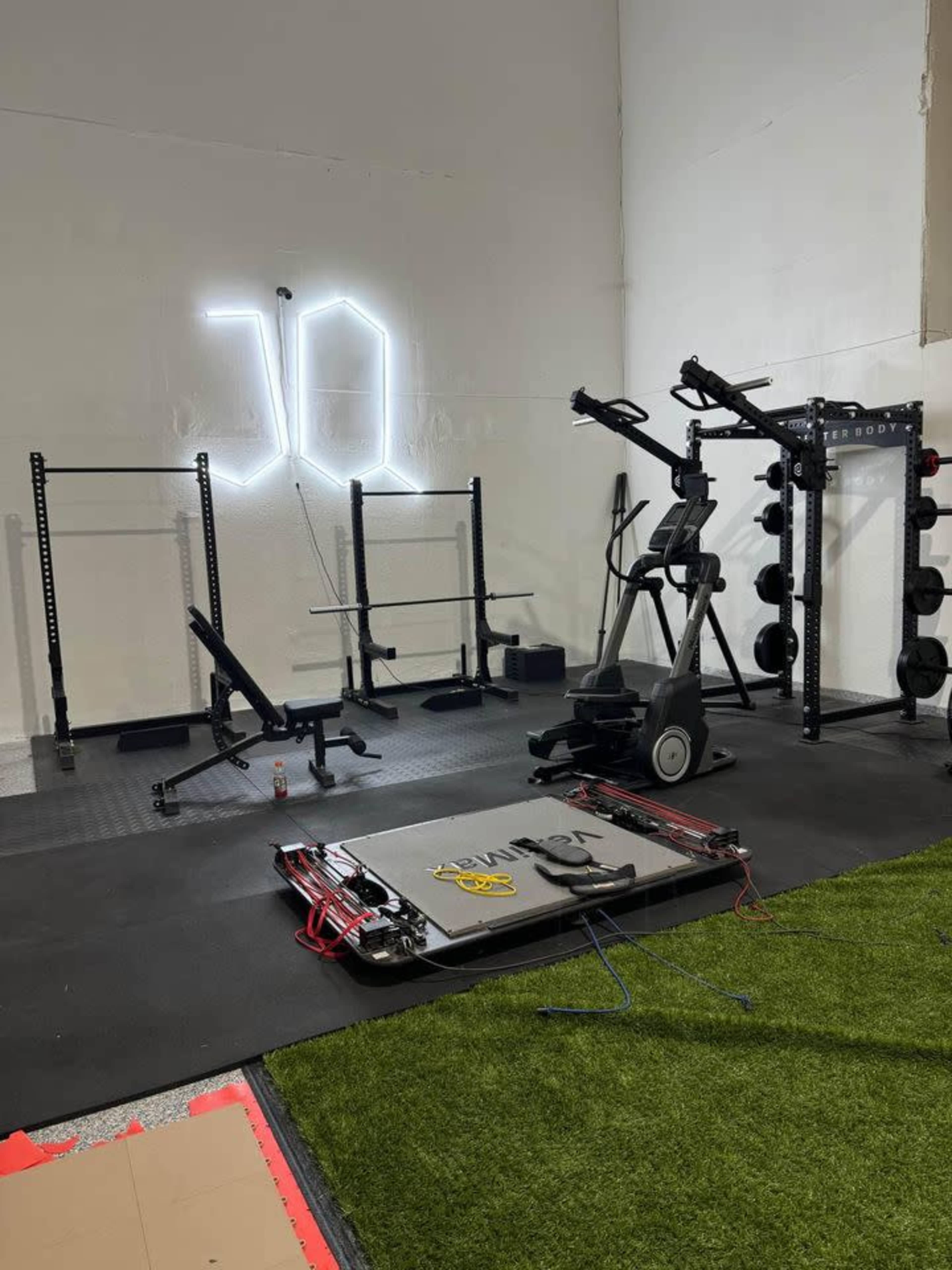 The image shows a workout space featuring various gym equipment, including weight racks, a multi-station machine, an elliptical, and free weights, all set against a bare wall with a neon light display.