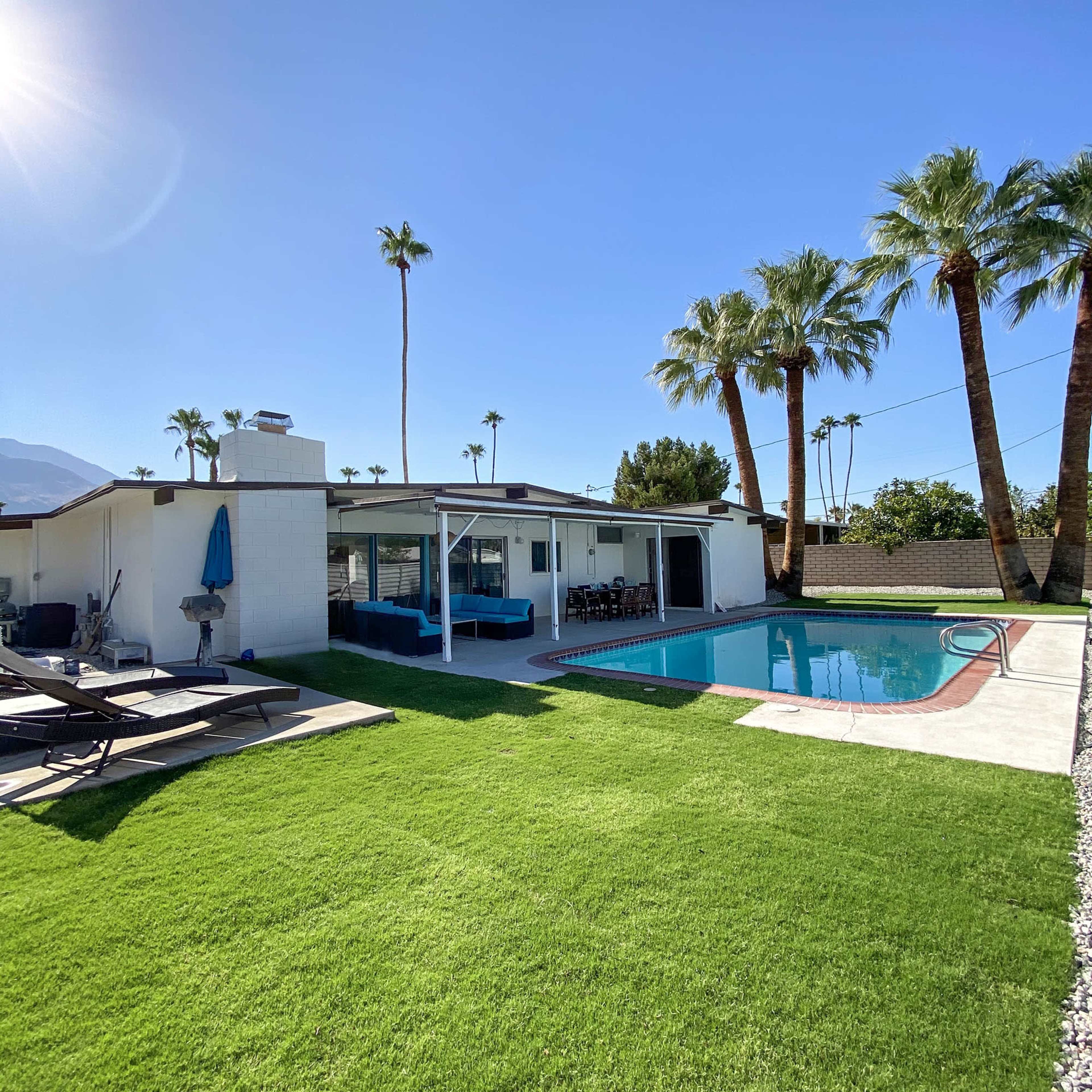 A modern house with a swimming pool is surrounded by green grass and palm trees.