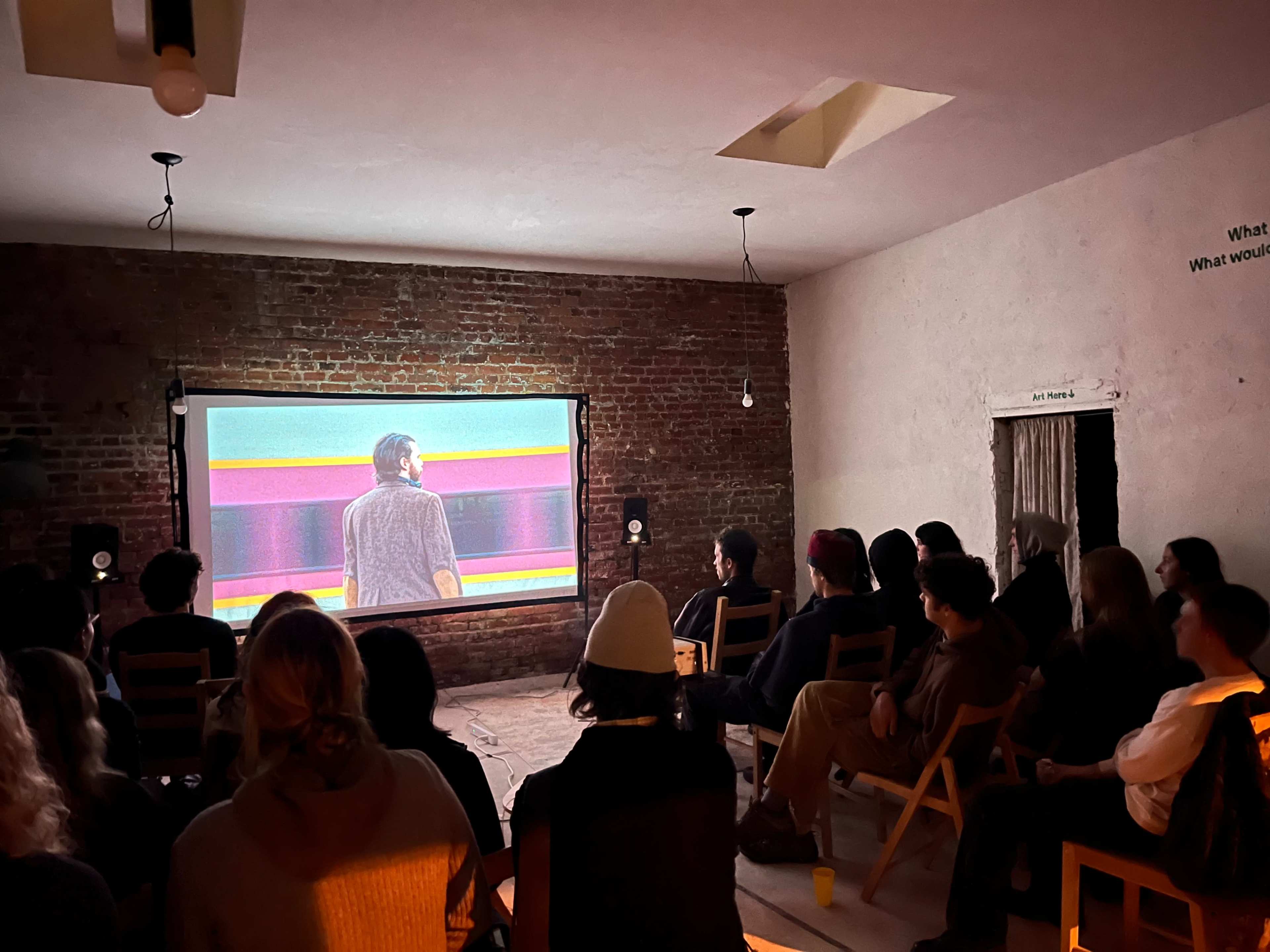 A group of people sits on folding chairs in a dimly lit room, watching a colorful film projected on a large screen.