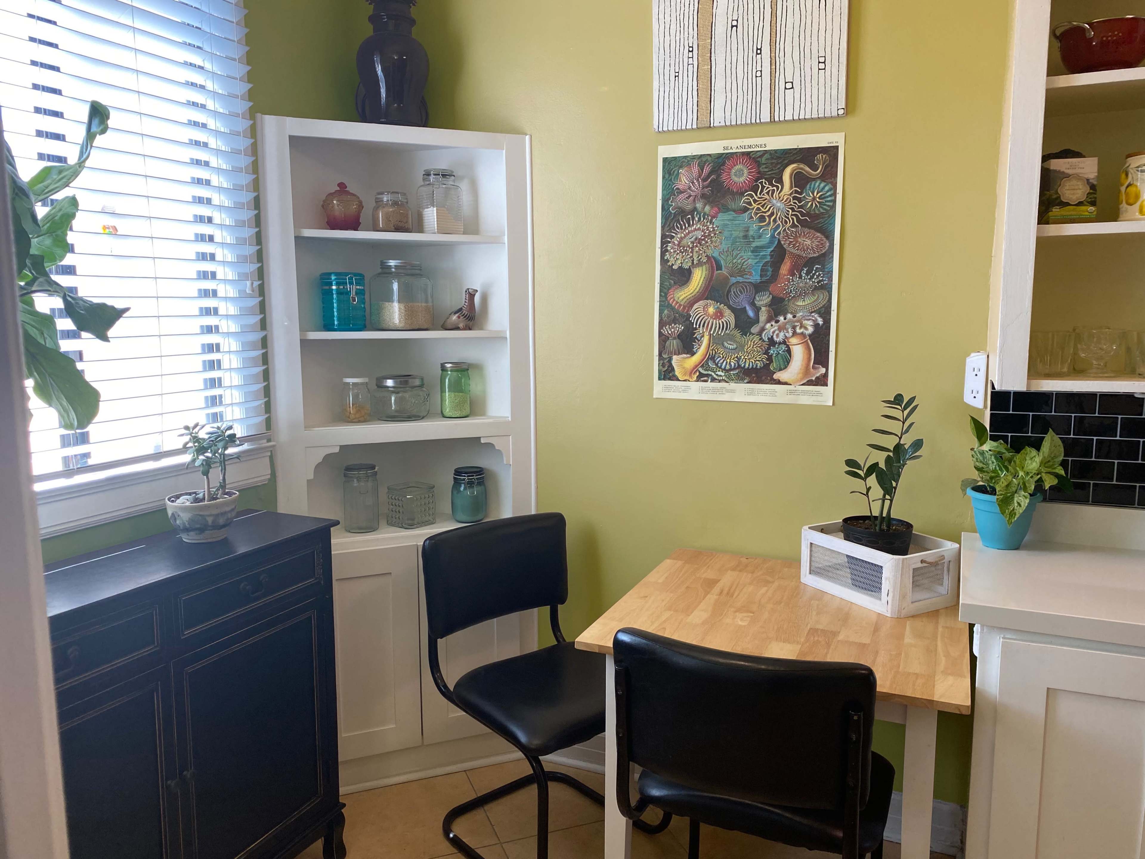 A small kitchen nook features a wooden table with two black chairs, a white shelving unit filled with jars and plants, and a colorful botanical poster on the wall.