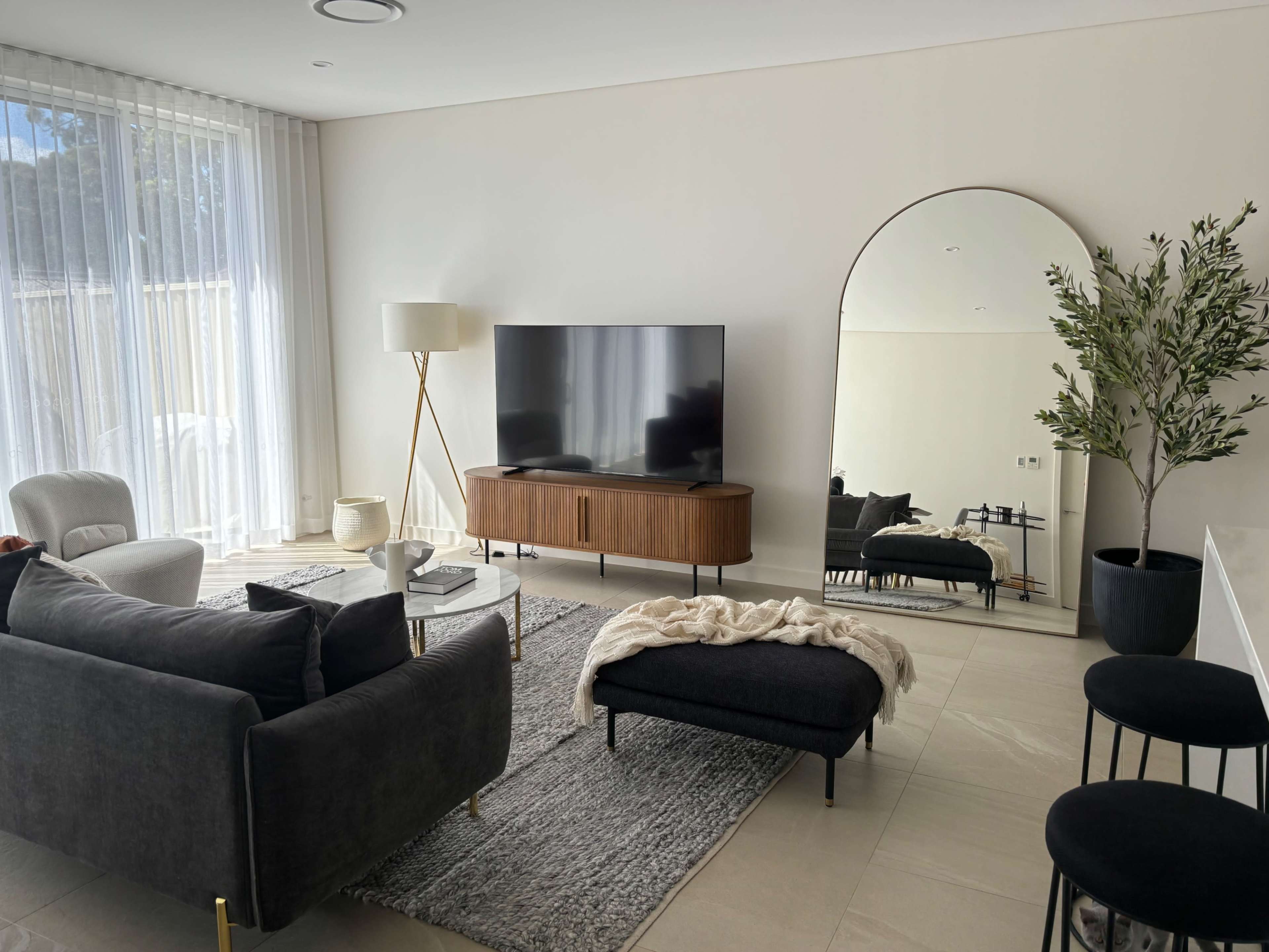 The living room features a dark sofa, a wooden TV cabinet, a large mirror, and a contemporary lamp, with light streaming through sheer curtains.