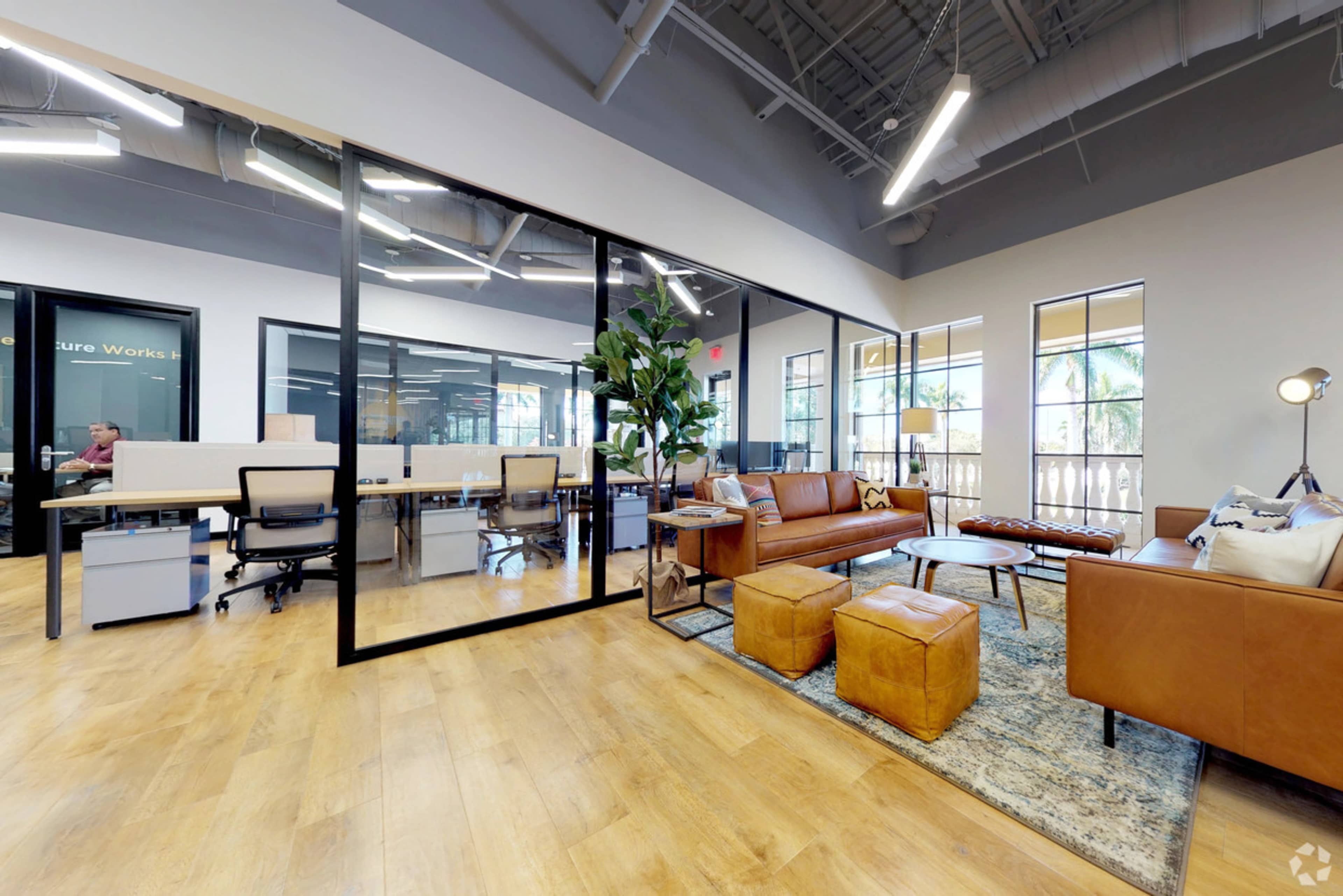 A modern office space featuring a lounge area with a brown leather couch, coffee table, and potted plant, adjacent to a workspace with desks and glass partitions.