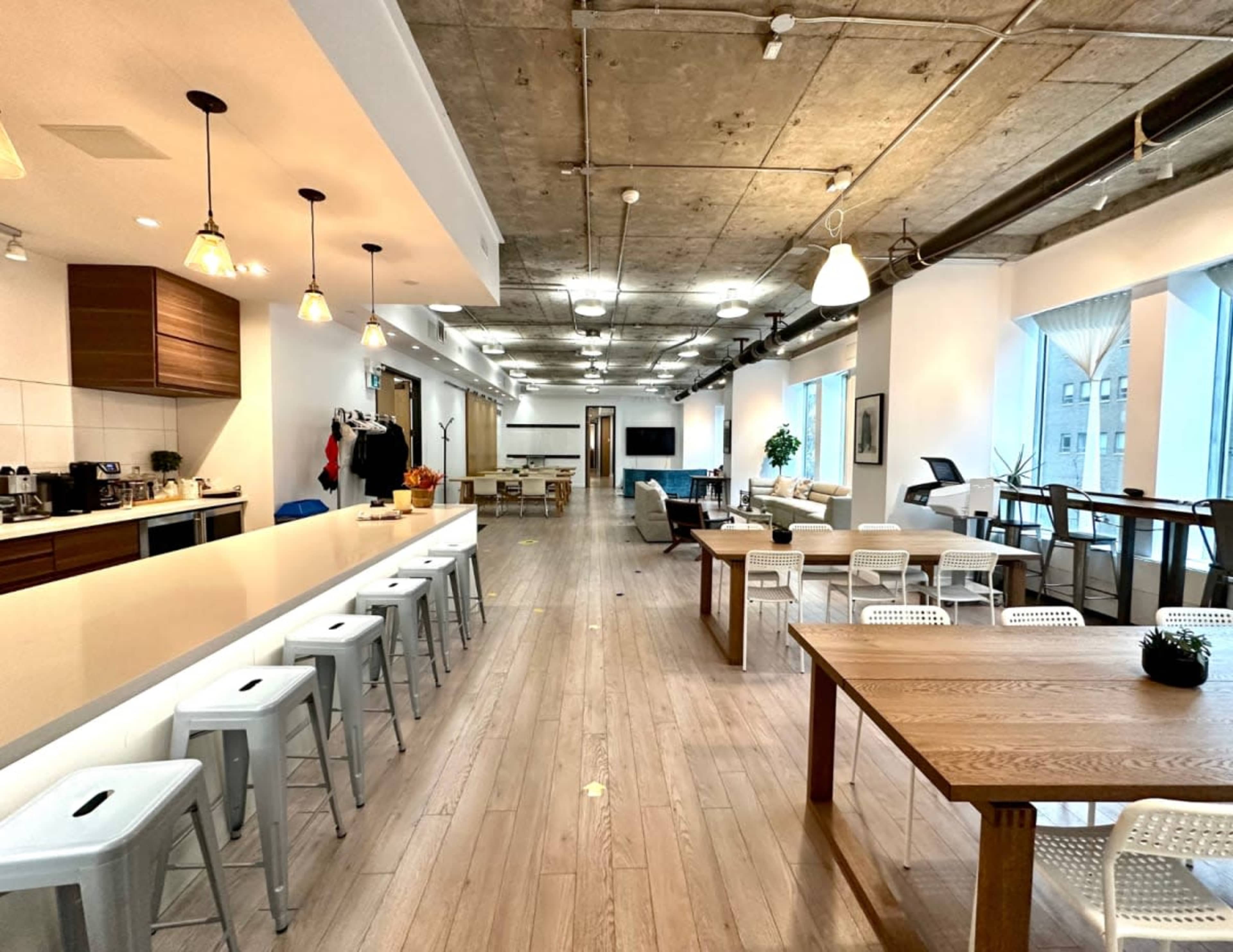 The image shows an open-concept modern office space with a long bar, seating areas, and large windows.