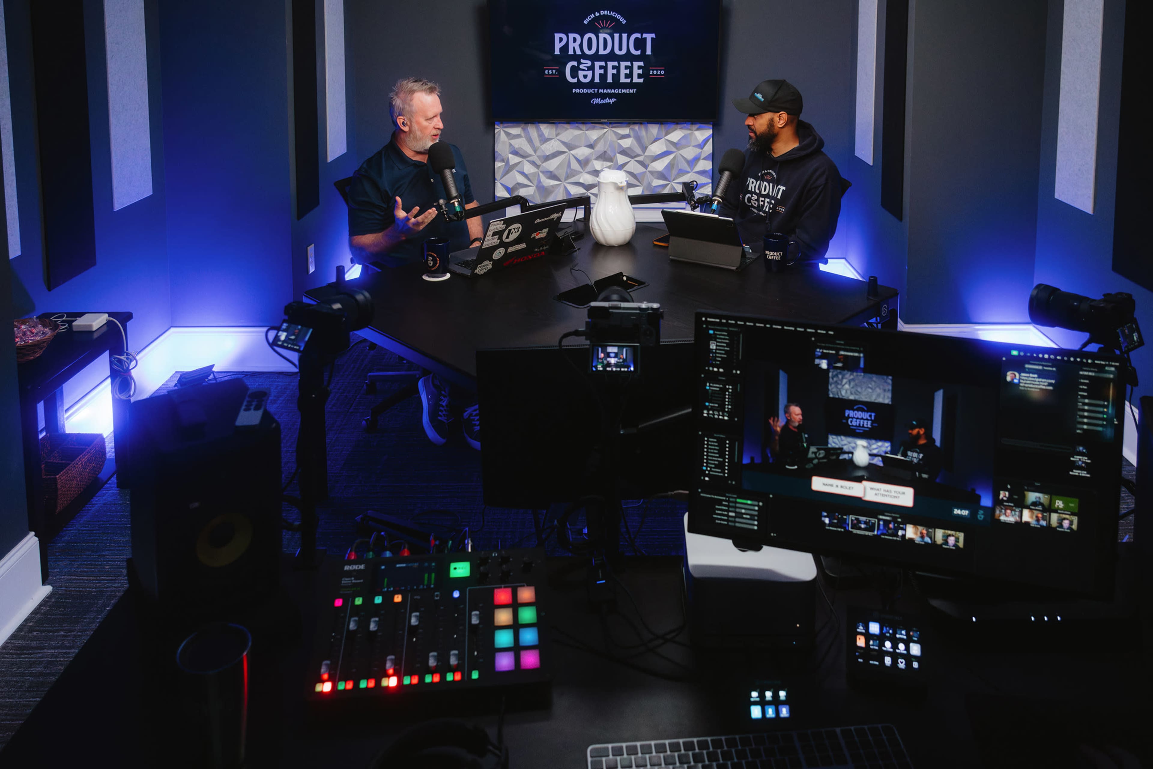 Two men are sitting at a modern conference table in a studio, discussing topics related to a show called "Product Coffee," with cameras and equipment set up around them.