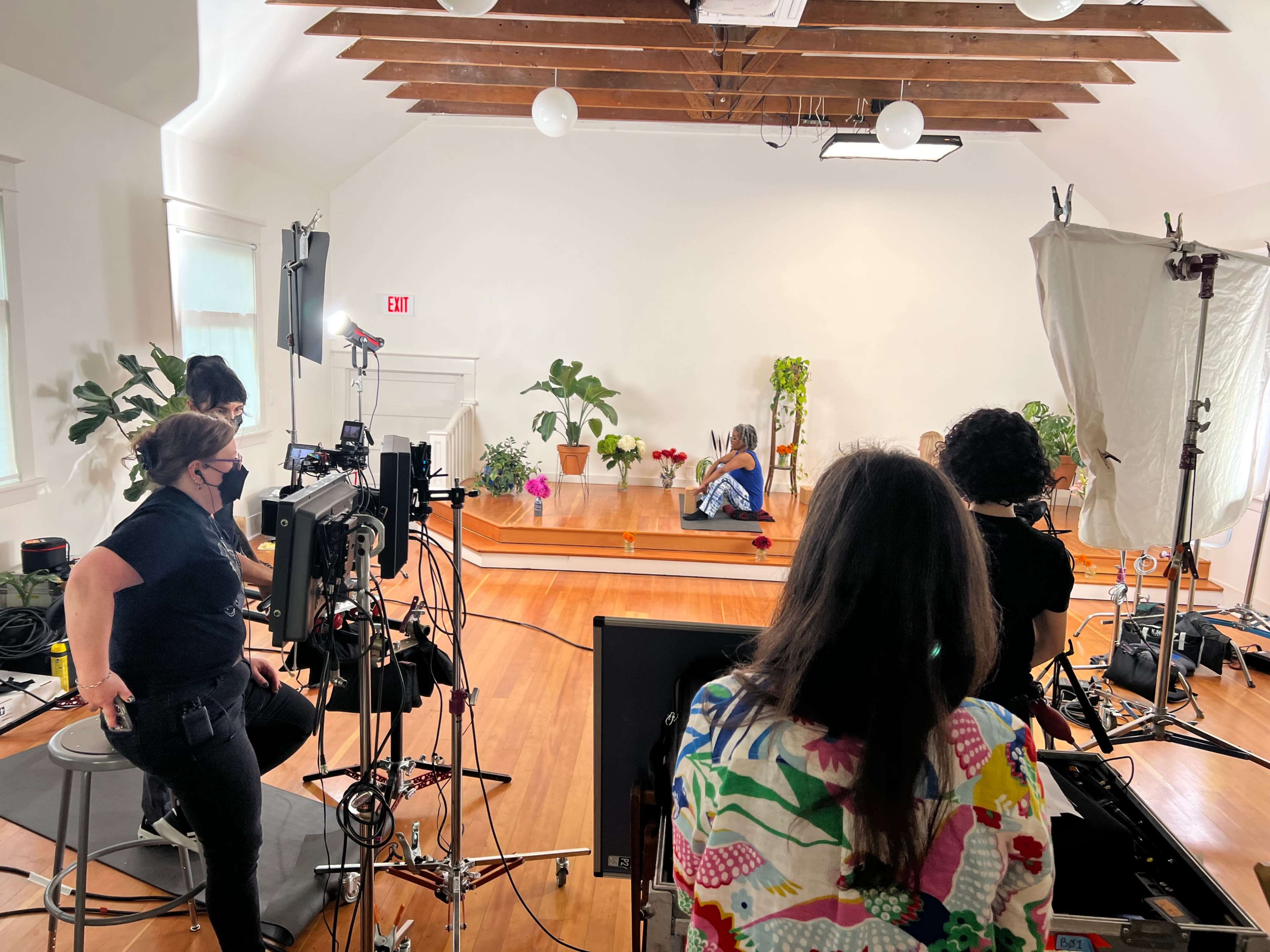 A filming setup in a bright room with wooden floors, where a crew is working around a person seated on a stage surrounded by plants and lighting equipment.