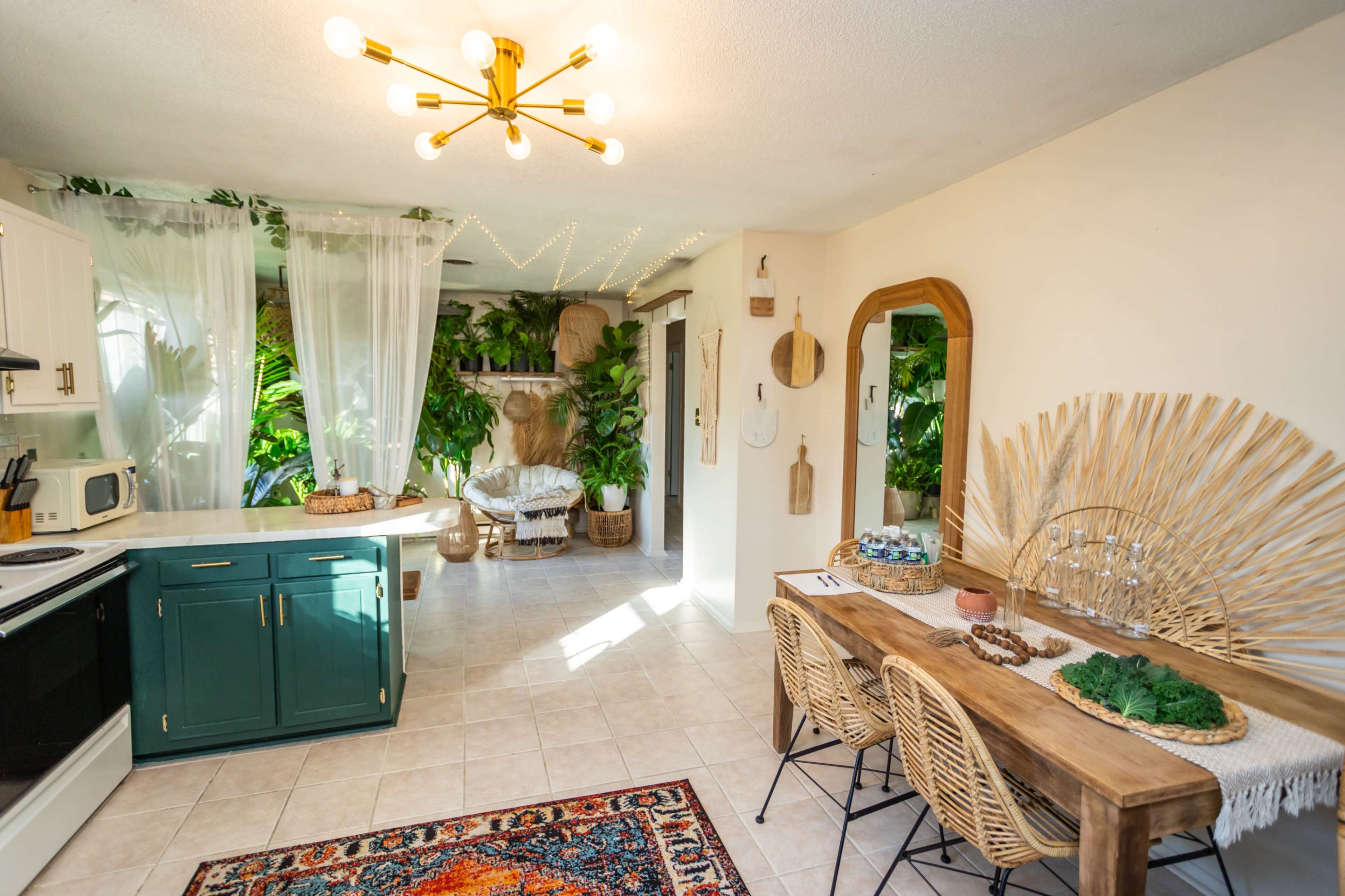 A spacious kitchen and dining area featuring a mix of modern and natural decor, with a large dining table, rattan chairs, and abundant greenery.