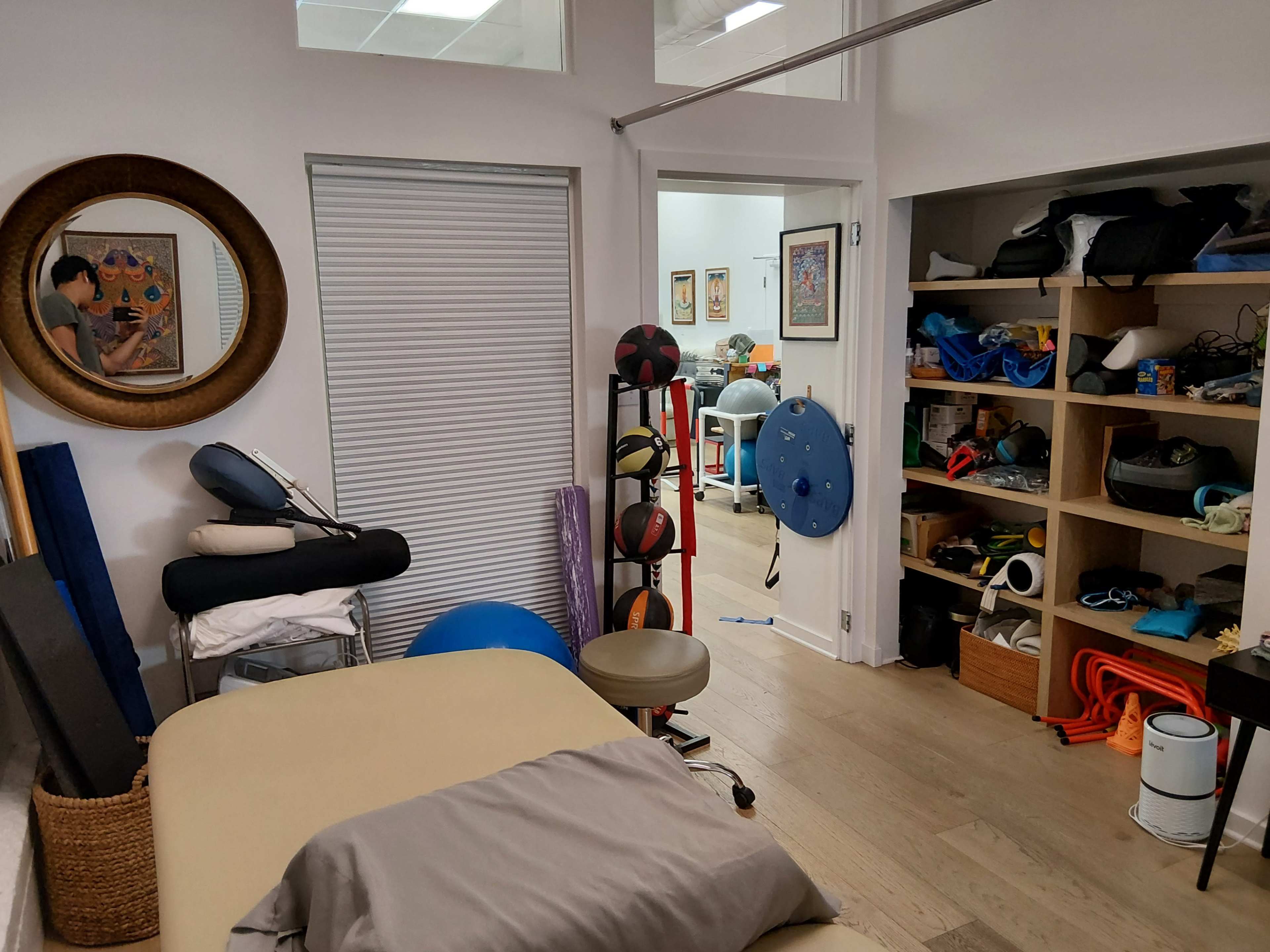 The image shows a small room with a massage table, exercise equipment, and shelving filled with various items.