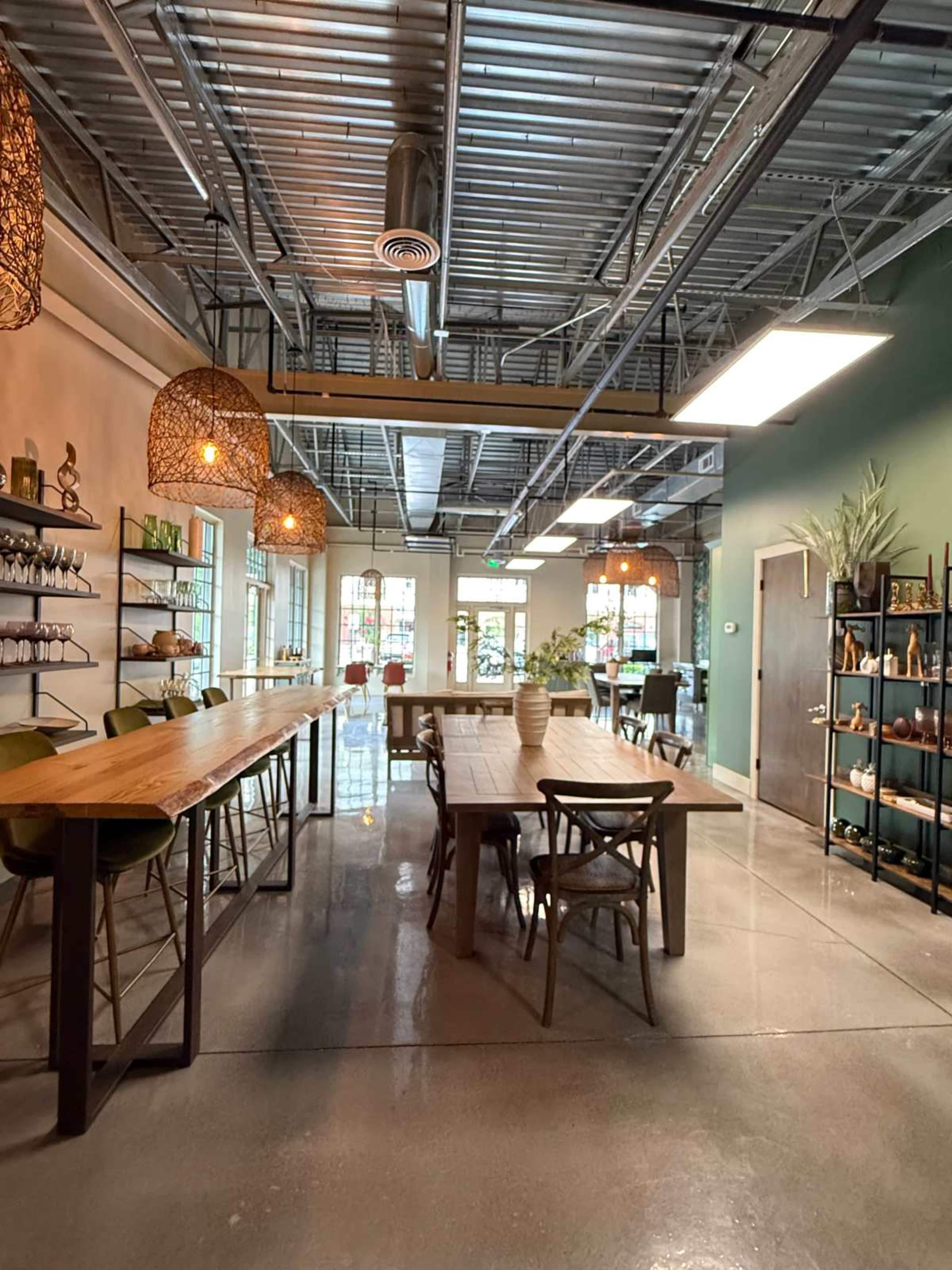 The image shows a modern, minimalistic dining area with wooden tables, metal shelves, and pendant lighting, featuring plants and decorative items spread throughout the space.