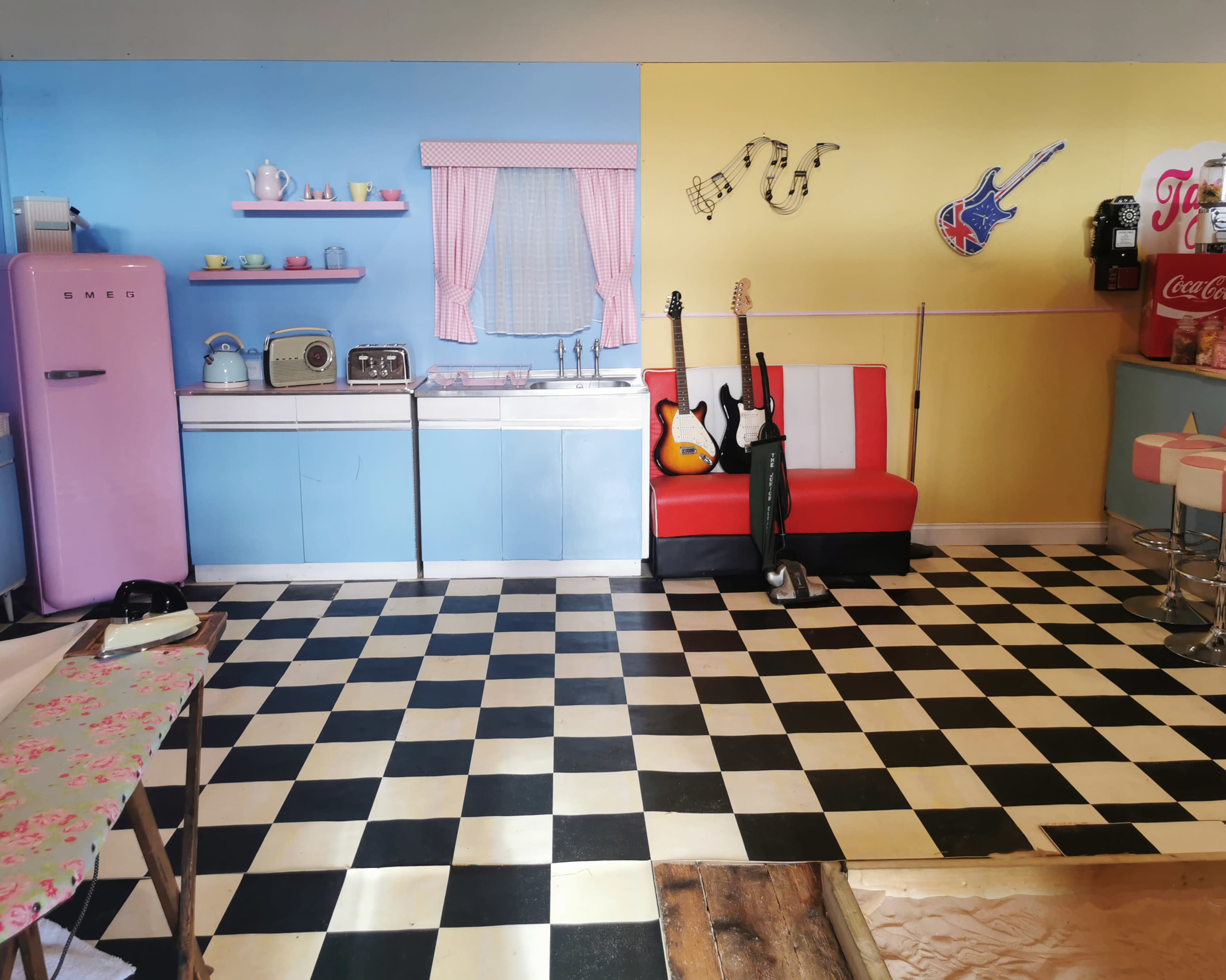 The image shows a vintage-style kitchen with a pink refrigerator, black-and-white checkered floor, and a red and white striped sofa, along with musical instruments and various kitchen appliances displayed.