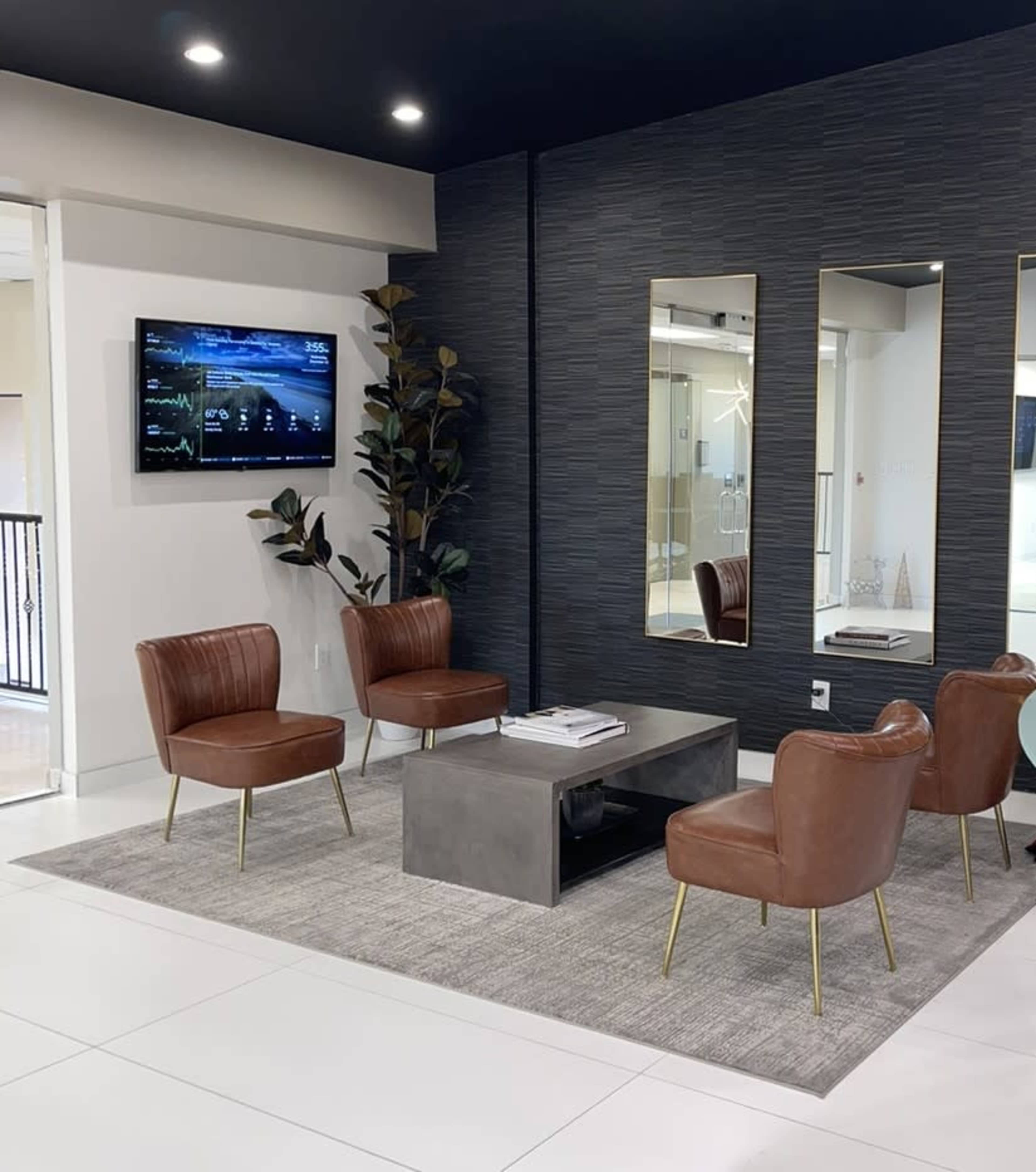 The image shows a modern waiting area featuring four brown leather chairs arranged around a metal table on a light gray carpet, with a television mounted on the wall and two mirrors flanking it.