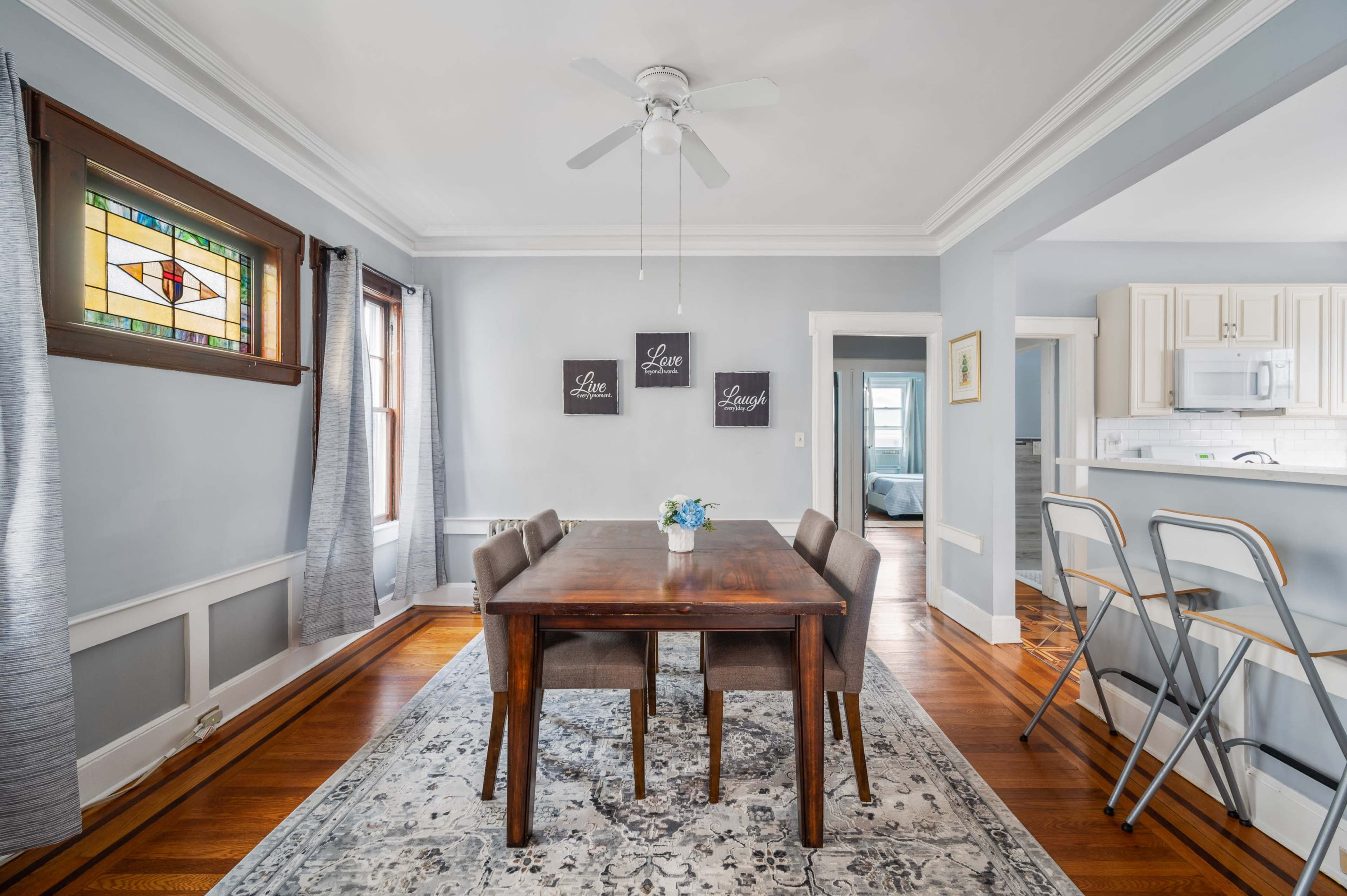 An airy dining room features a wooden table surrounded by chairs, with a stained glass window and artwork on the walls.