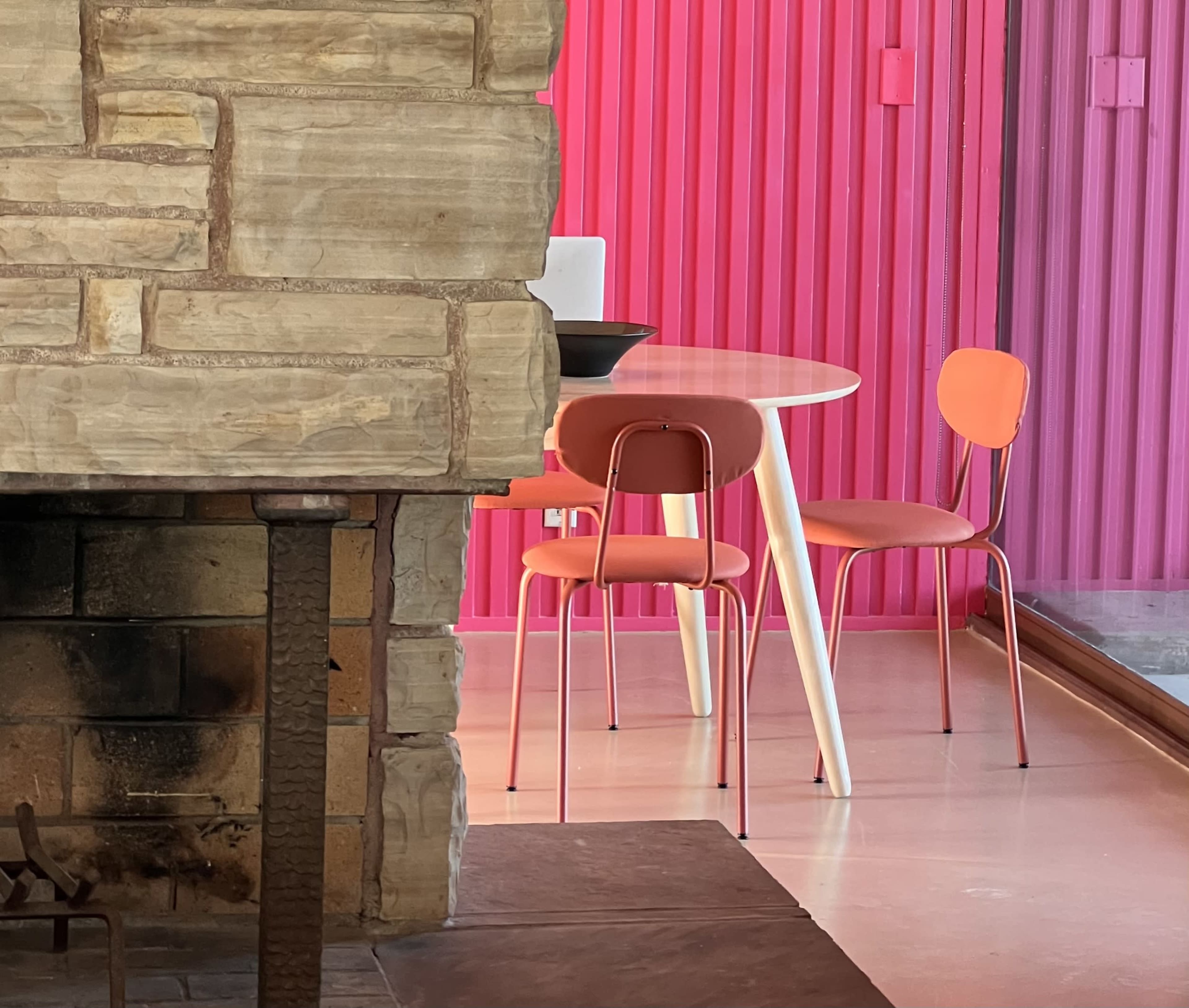 A stone fireplace is in the foreground, with a round table and three chairs in vibrant colors set against a pink paneled wall in the background.