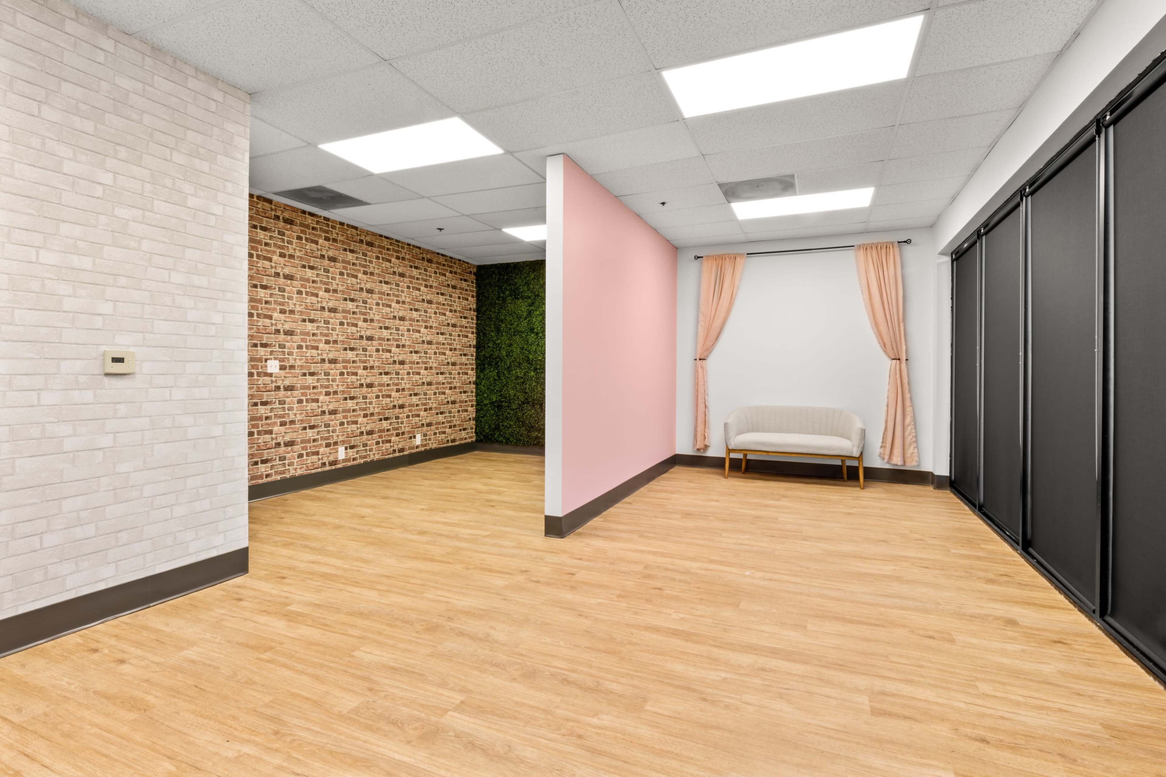 The image shows a spacious room with a combination of textured walls, including exposed brick, a solid pink wall, a green plant wall, and a light wood floor.
