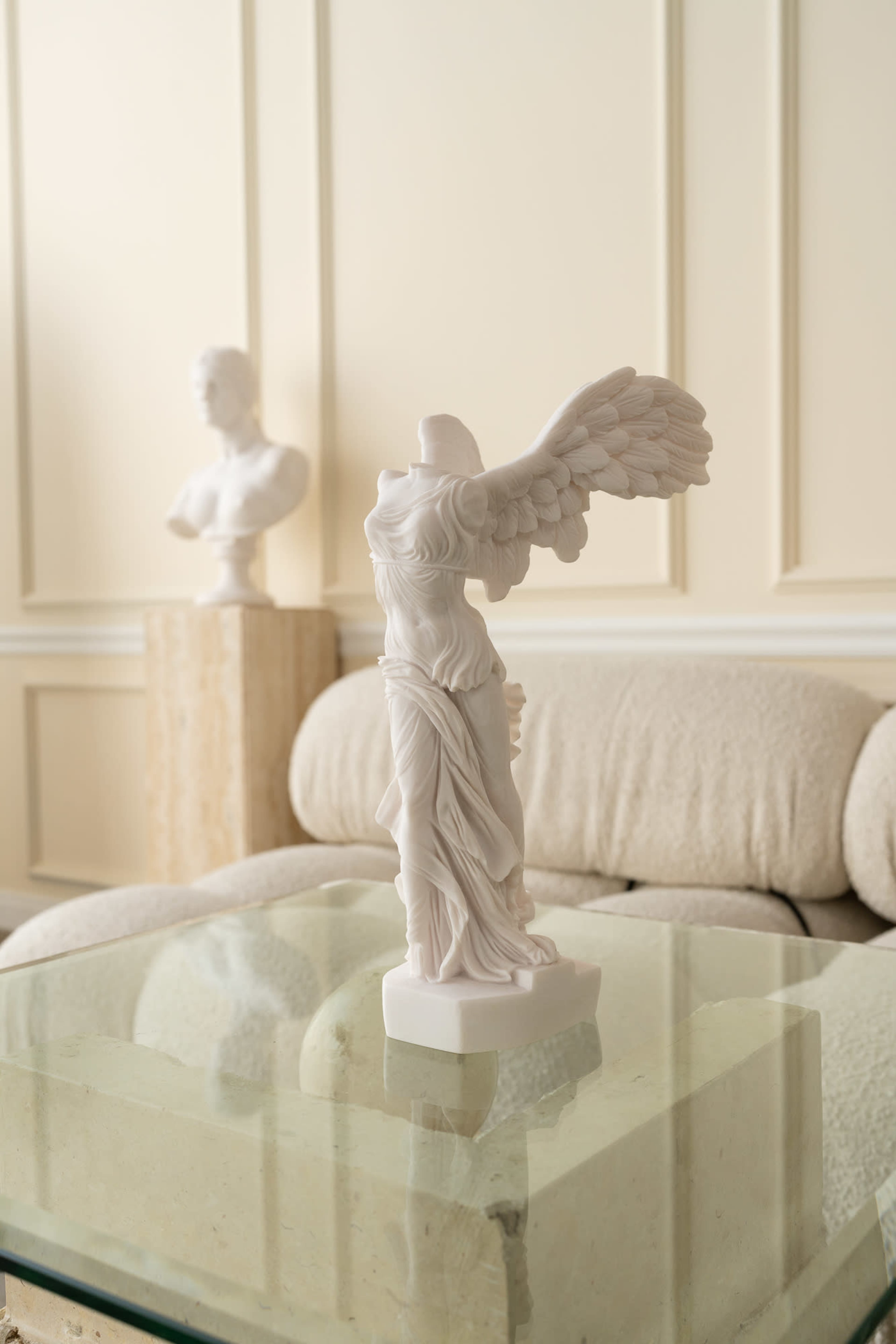 A marble sculpture of the Winged Victory of Samothrace is displayed on a glass table in a room with light-colored walls and a sofa.