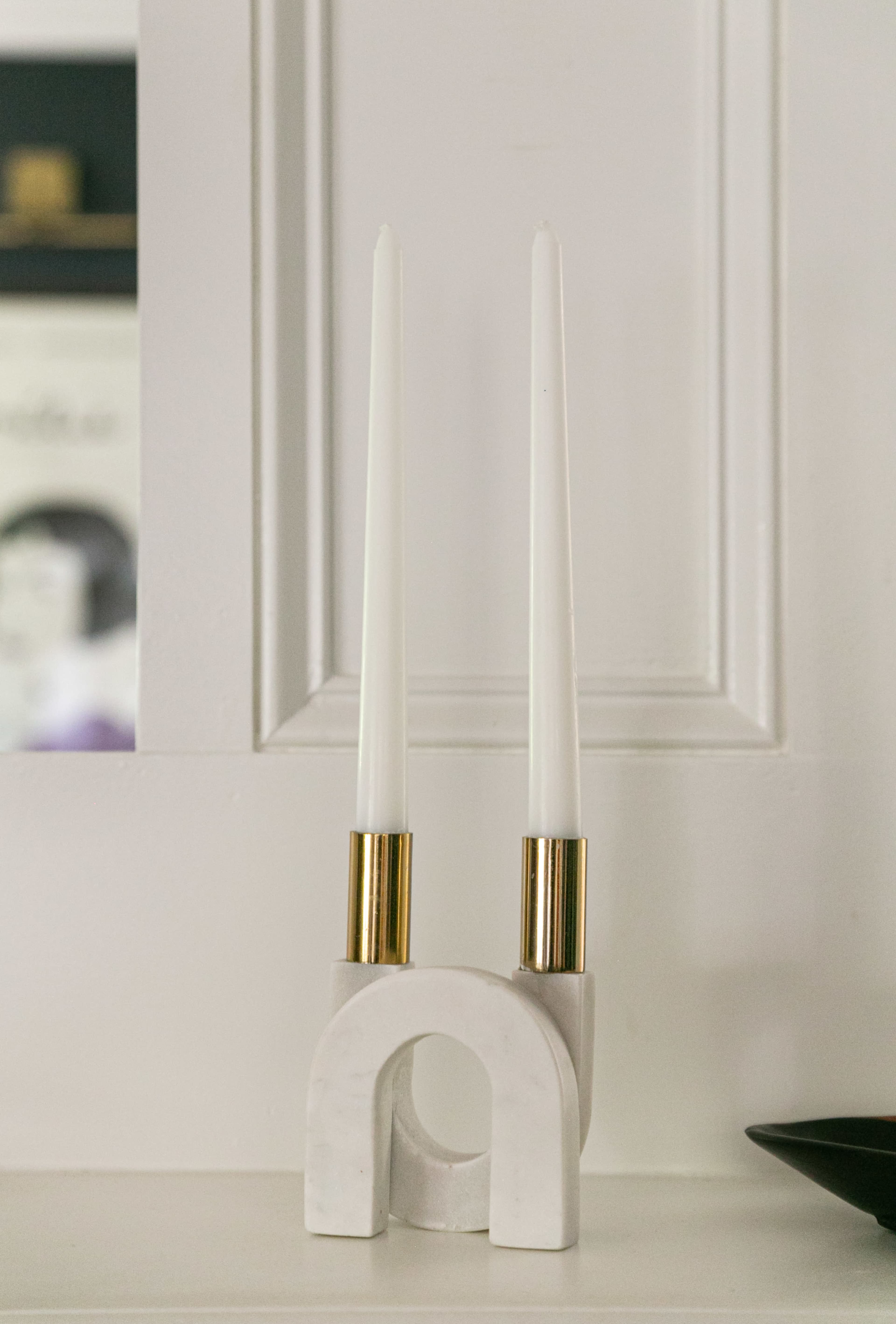Two white candles are placed in a marble candle holder on a shelf against a white wall.