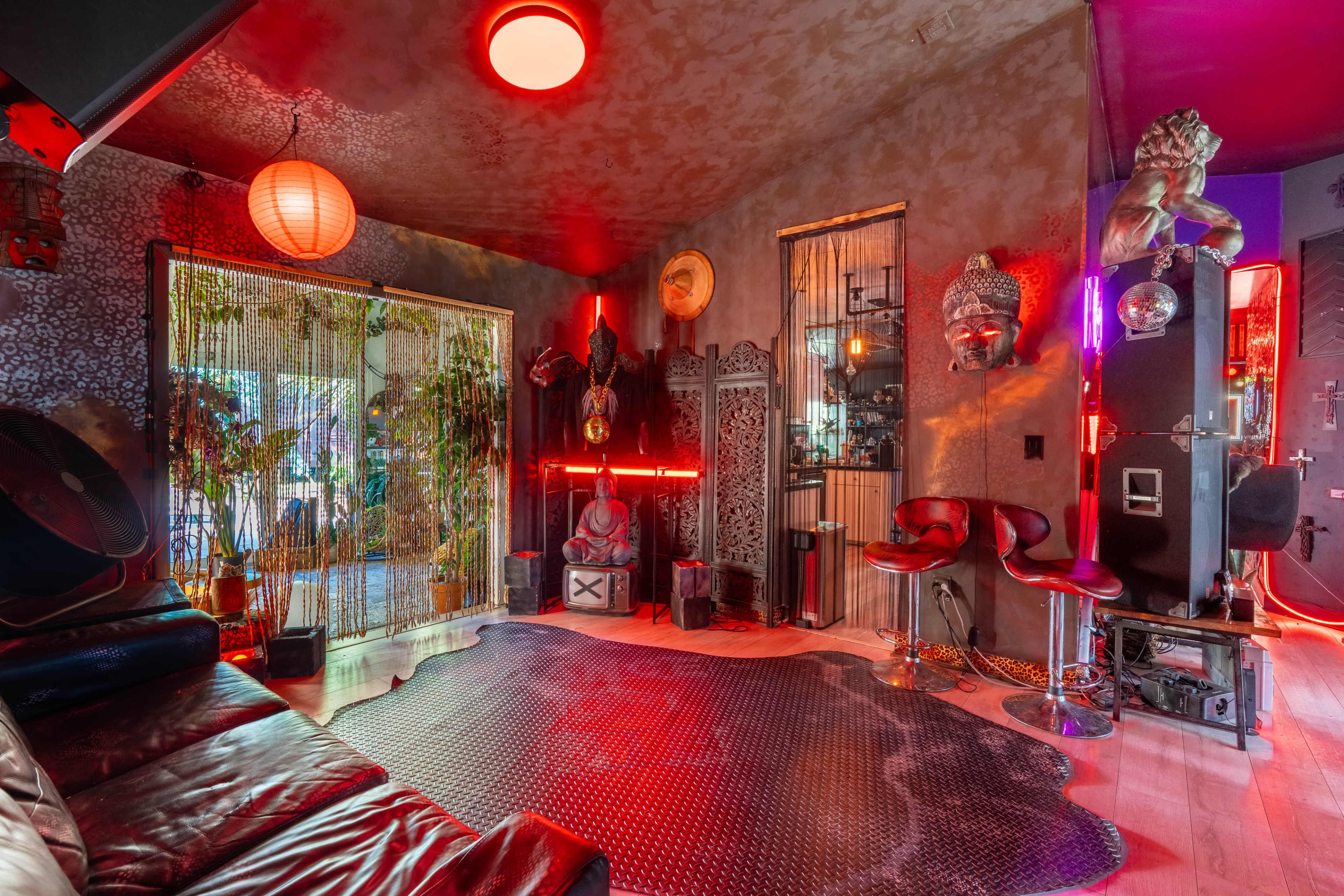 The image shows a dimly lit room with eclectic decor, featuring a red and black color scheme, various ornamental statues, and modern furniture arranged around a unique patterned floor.