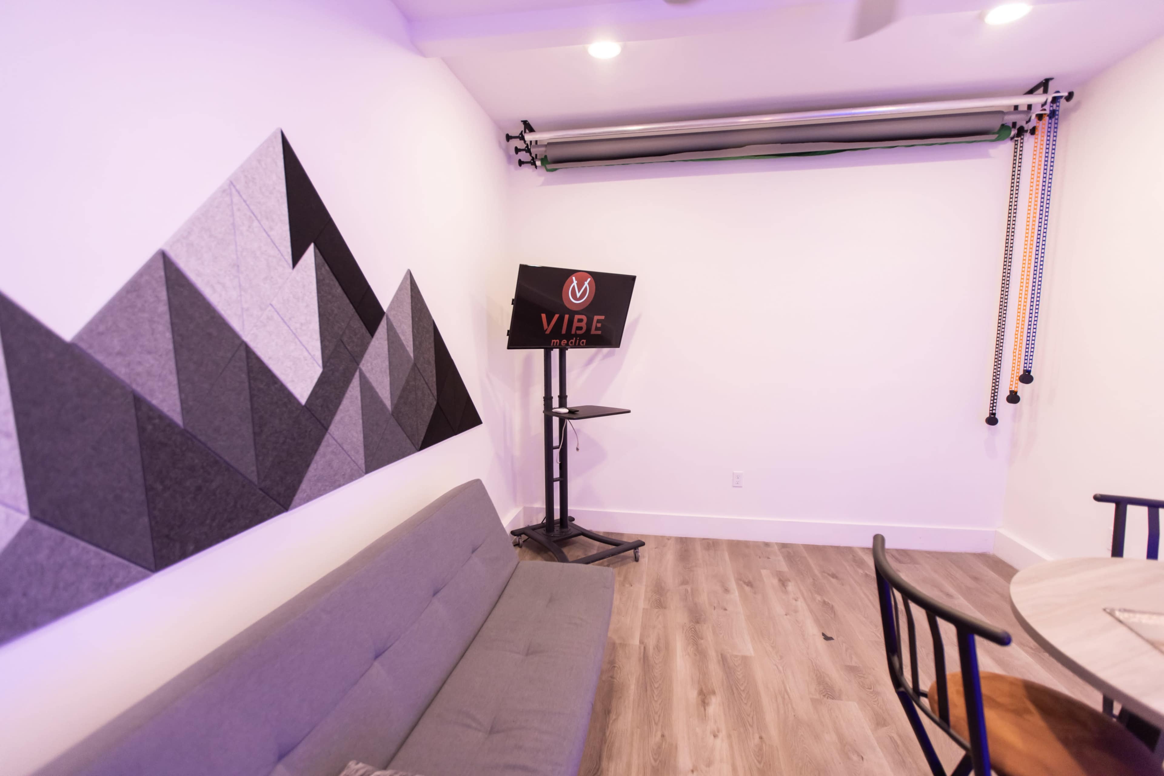 The image shows a minimalist room featuring a gray sofa, a mountain-themed wall decoration, and a VIBE Pro display stand in the corner.