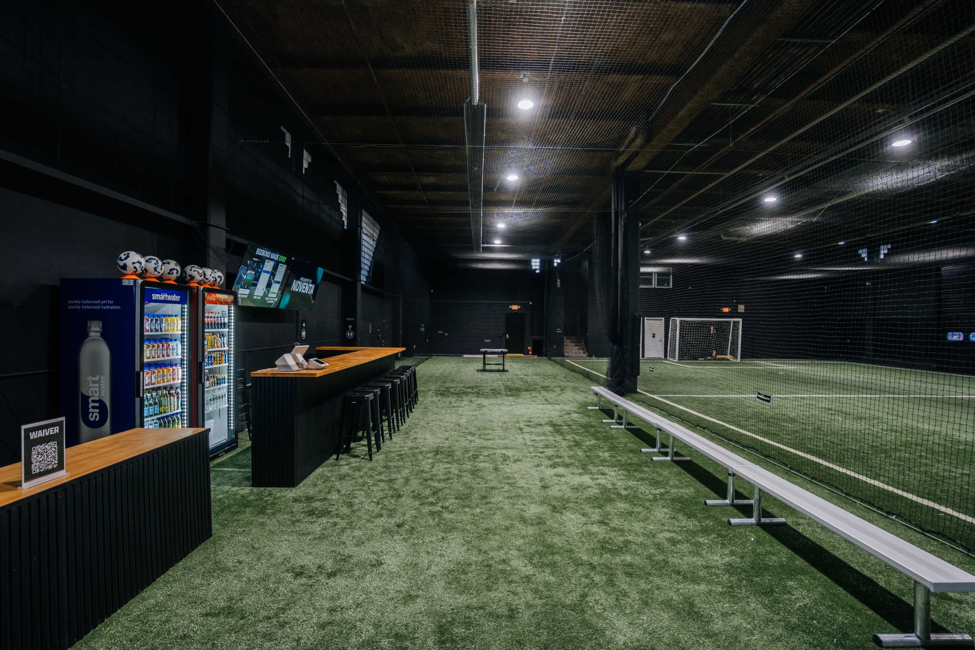 The image shows an indoor sports facility with artificial turf, a concession area with vending machines, and a soccer goal in the distance.