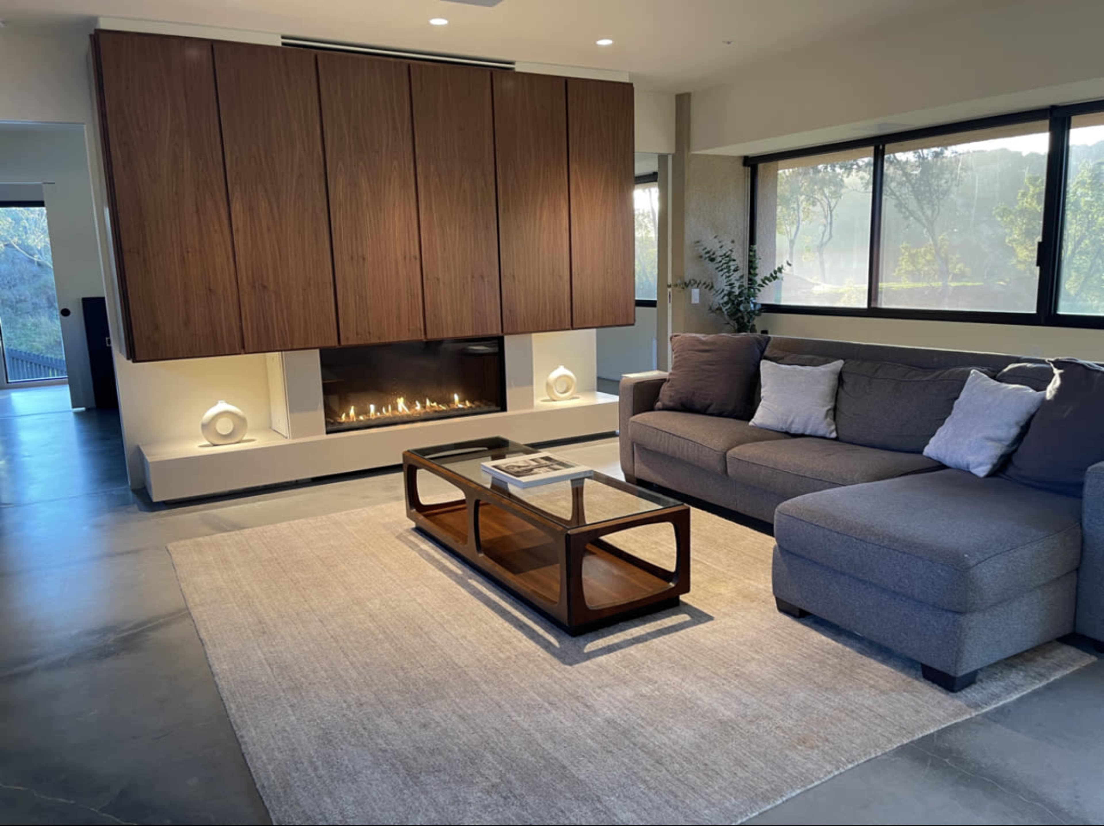 A modern living room features a gray sectional sofa, a glass coffee table, and a built-in fireplace with a wood panel backdrop.