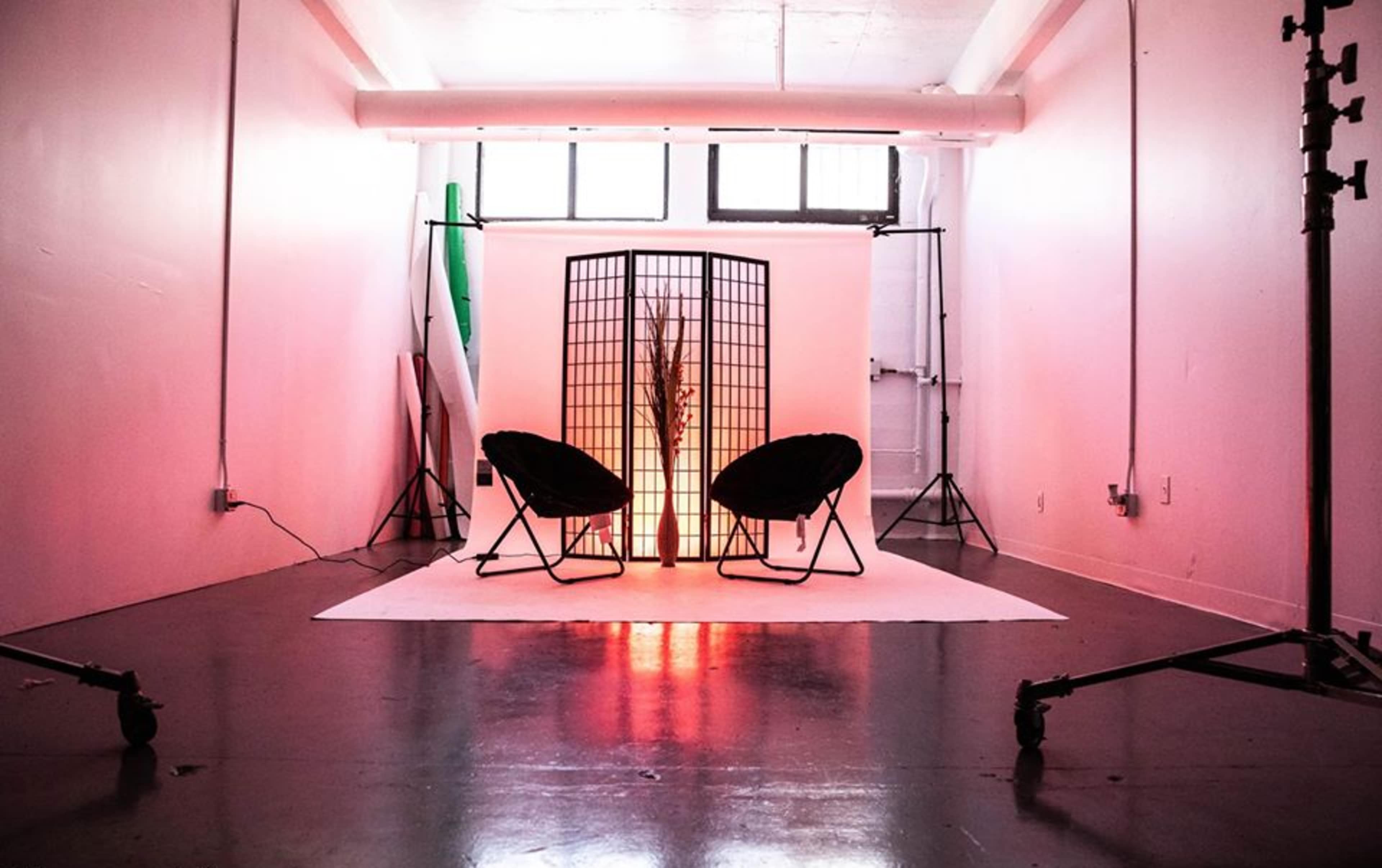 A studio setup featuring two chairs facing a decorative screen, illuminated by soft light, with a backdrop of various photography equipment and colored backgrounds.
