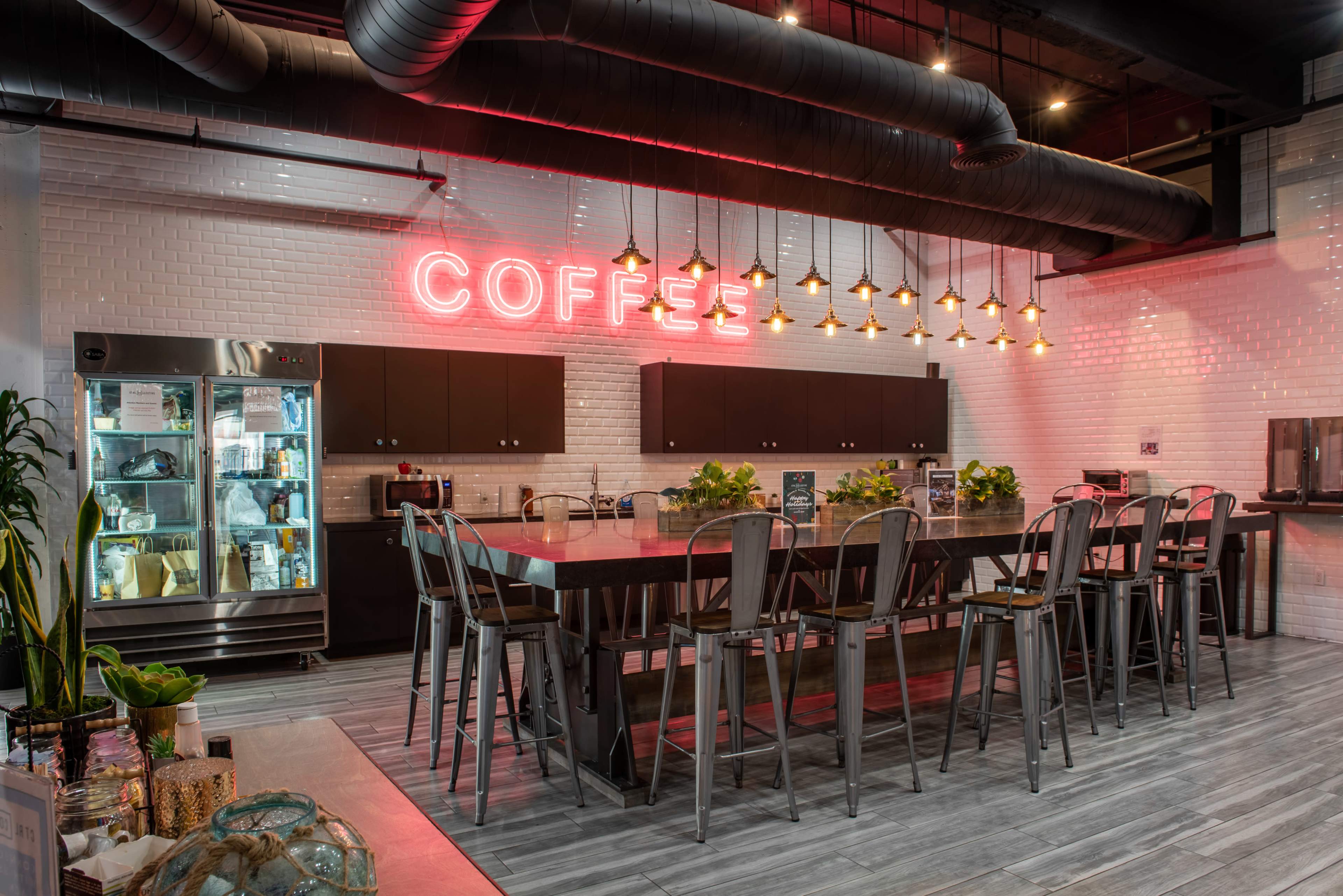 A modern coffee bar features a long table with metallic stools, illuminated by a neon sign reading "COFFEE" and surrounded by sleek cabinetry and a glass refrigerator.