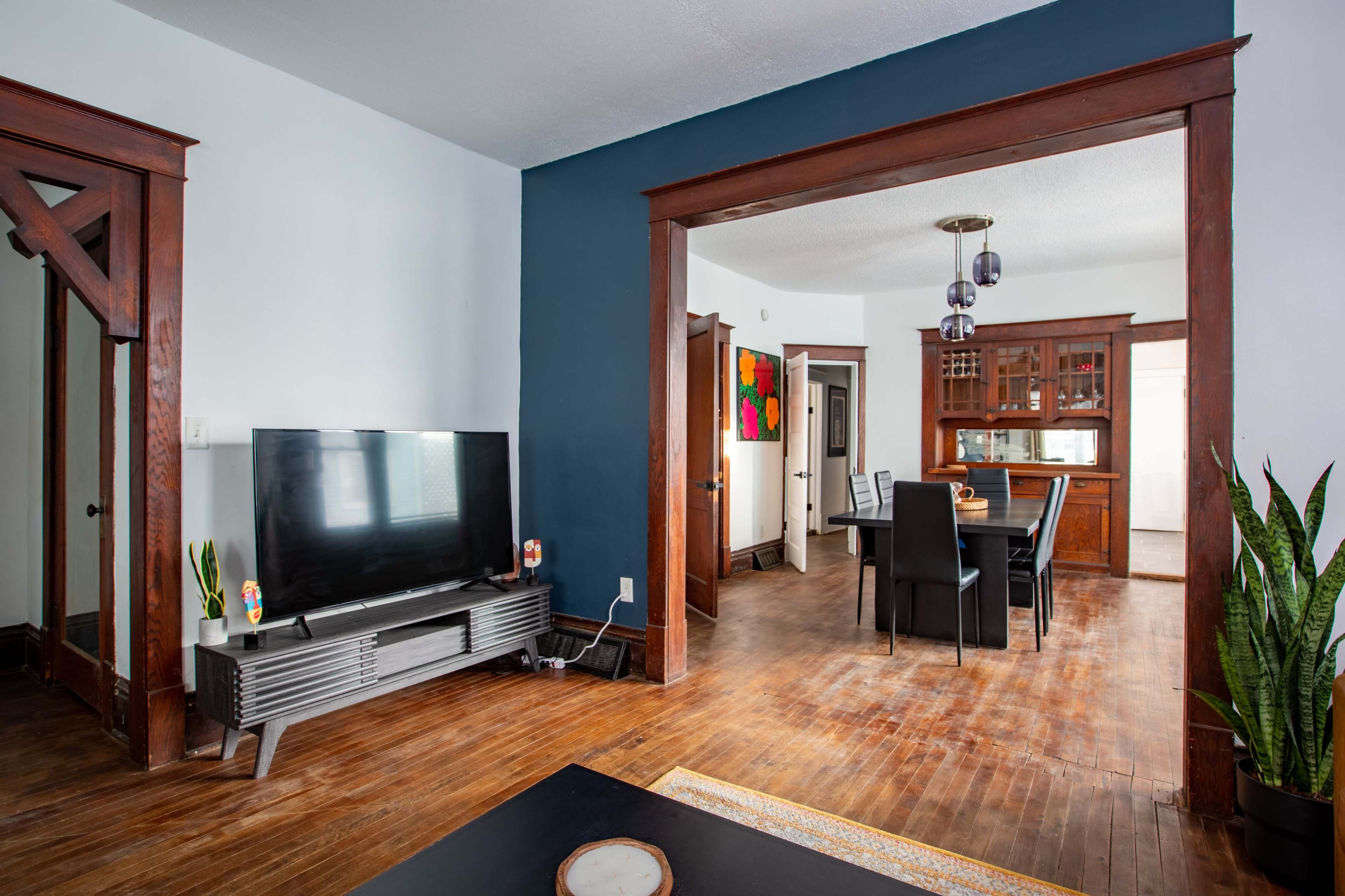 The image shows a living room with a television on a stand, hardwood floors, and a dining area visible in the background with a table and chairs.