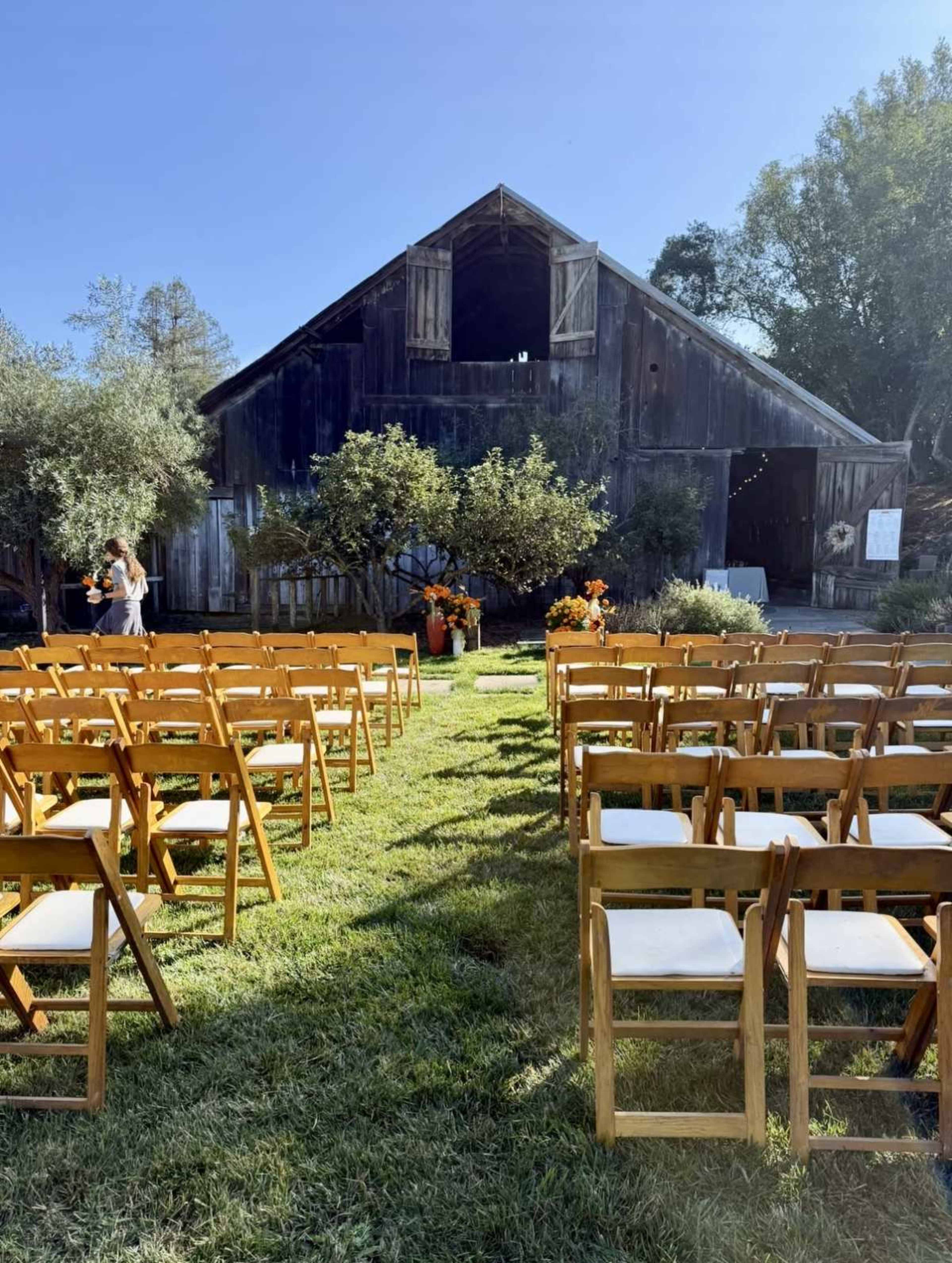 A rustic barn serves as a backdrop for a wedding ceremony setup, with rows of wooden chairs arranged on a grassy area and floral decorations nearby.