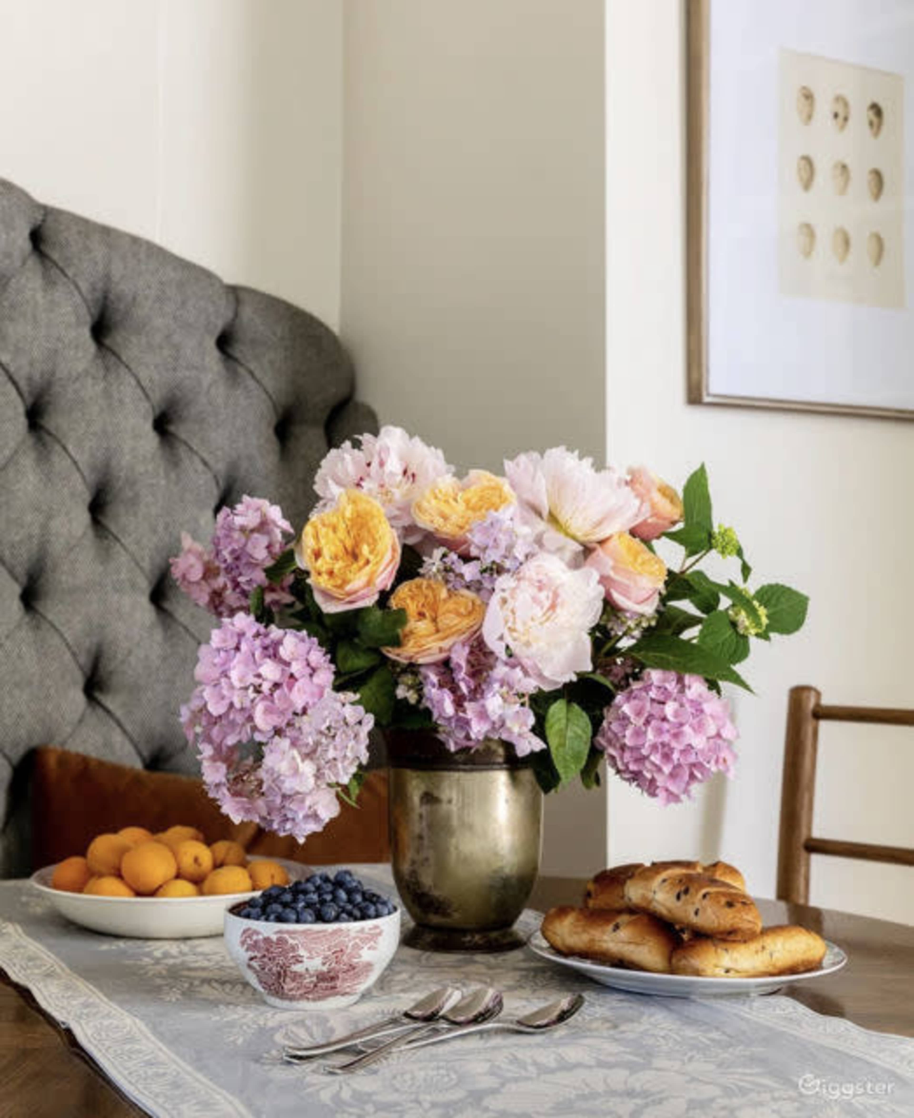 A table is set with a floral arrangement in a brass vase, a bowl of apricots, a bowl of blueberries, and a platter of bagels on a lace tablecloth.