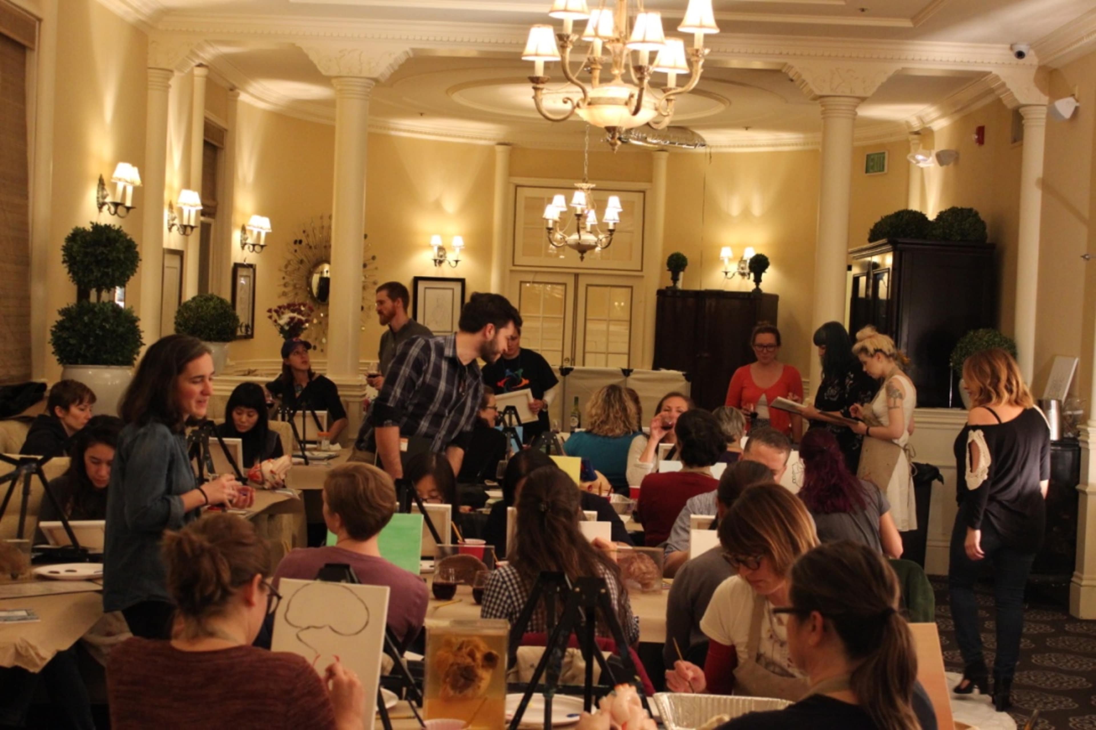 A group of people participates in a painting class in an elegantly decorated room with tables, easels, and art supplies.