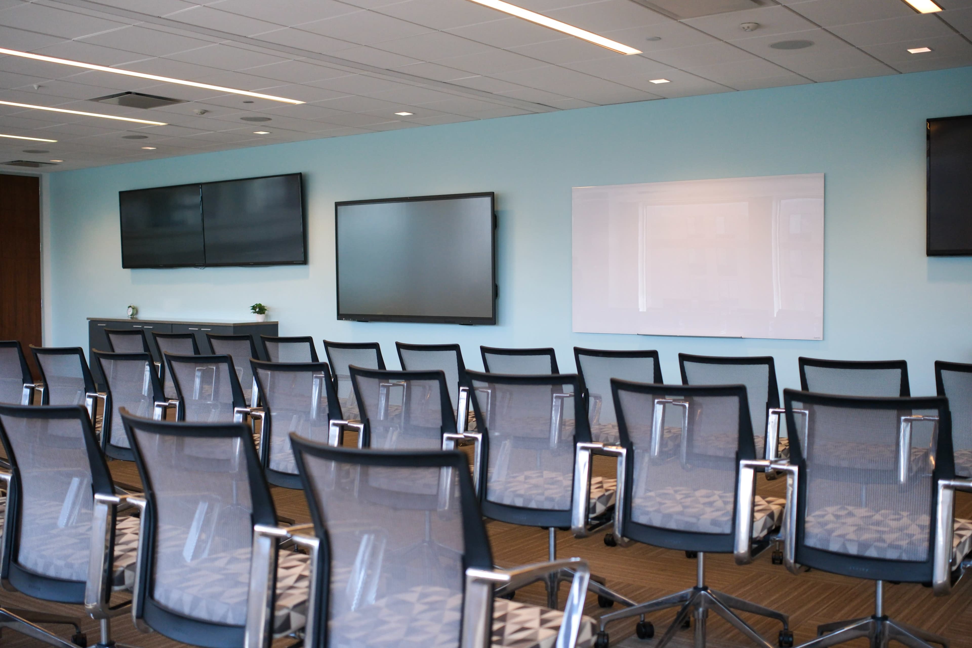 The image shows a modern conference room with several rows of mesh office chairs facing two wall-mounted screens and a whiteboard.