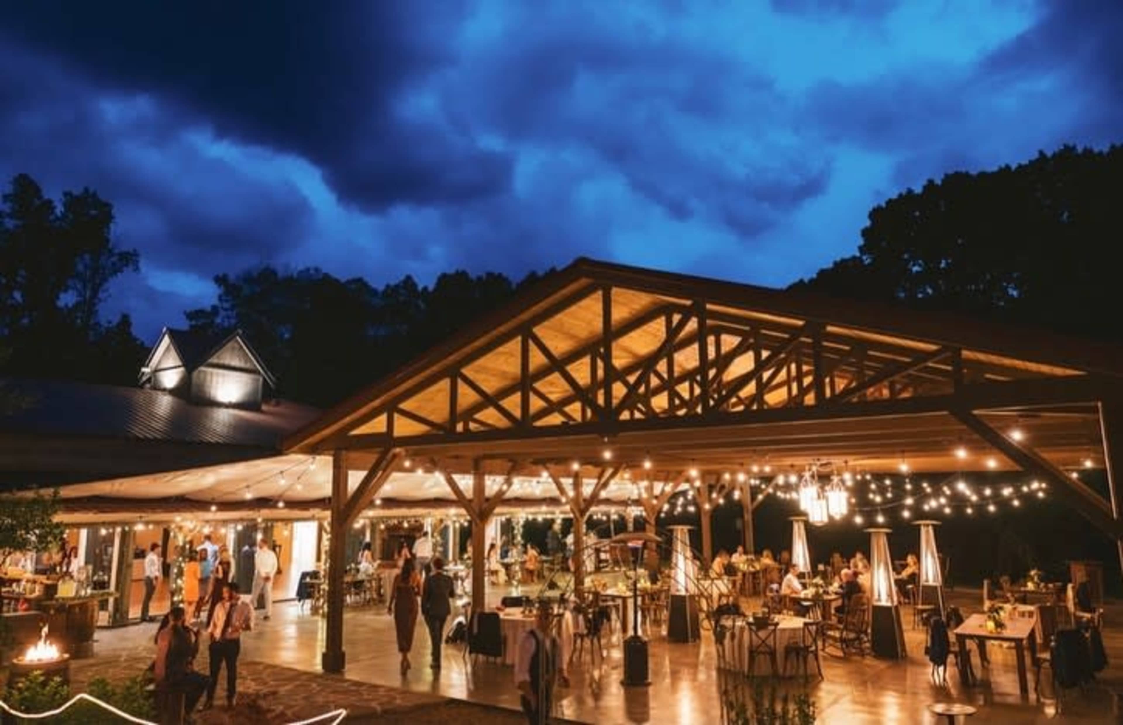 A spacious outdoor venue with a wooden structure is illuminated by string lights, hosting guests seated at tables under a darkening sky.